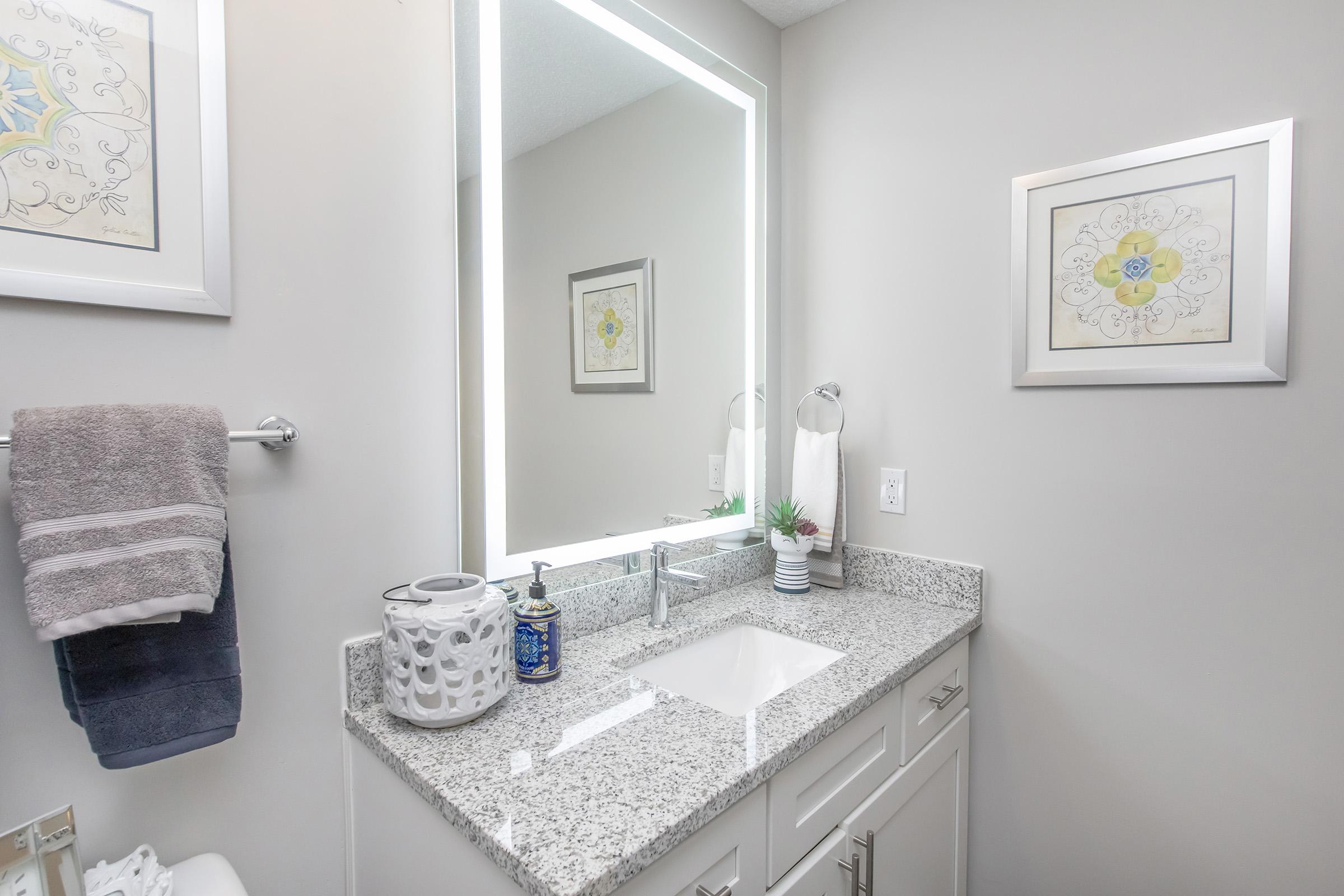 A modern bathroom featuring a white sink with a granite countertop, illuminated mirror, and decorative elements. There are towels hanging on a towel bar, a decorative candle holder, and framed art on the wall, creating a clean and stylish ambiance.