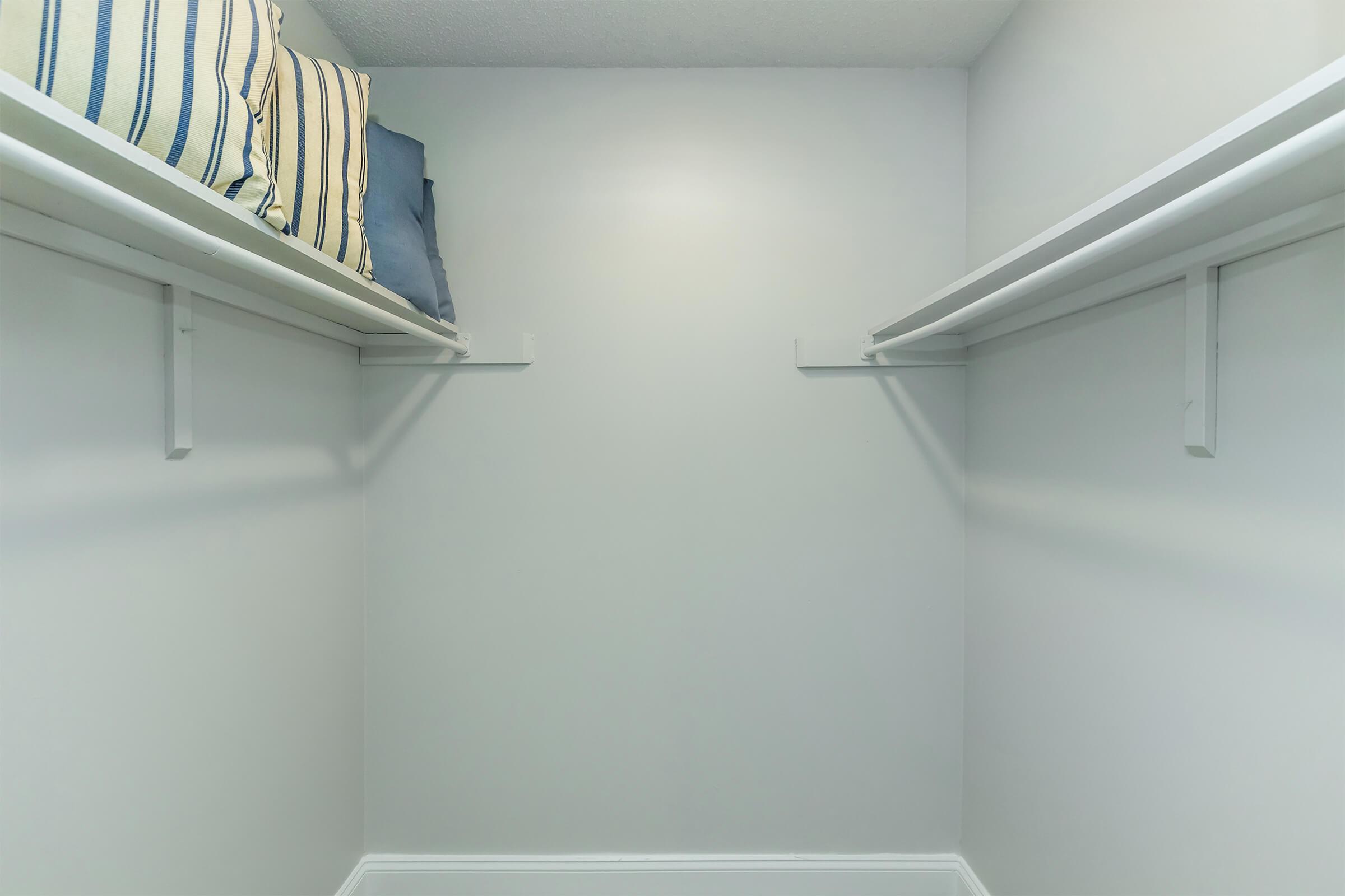 An empty closet featuring light gray walls, two white shelving units on either side, and neatly stacked pillows with blue and striped covers on the left shelf. The space appears clean and well-organized, providing a simple and minimalistic storage area.