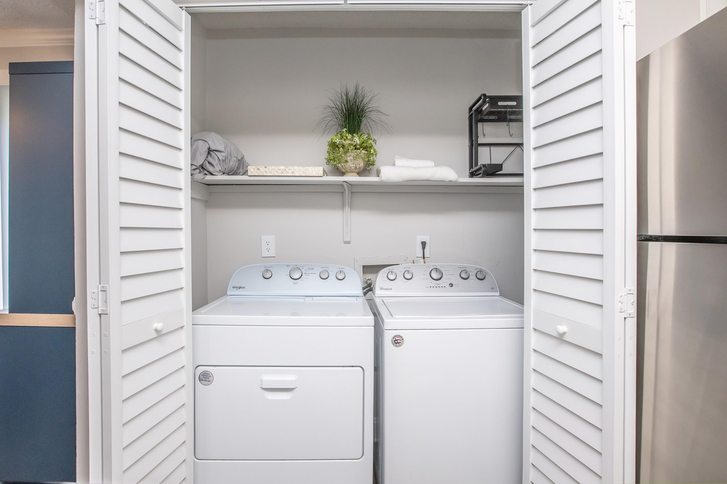 A laundry closet featuring a stacked washer and dryer. Above them, a shelf holds neatly folded towels and a small potted plant. The doors are louvered and closed, creating a tidy and organized appearance, with a stainless steel refrigerator visible to the side.