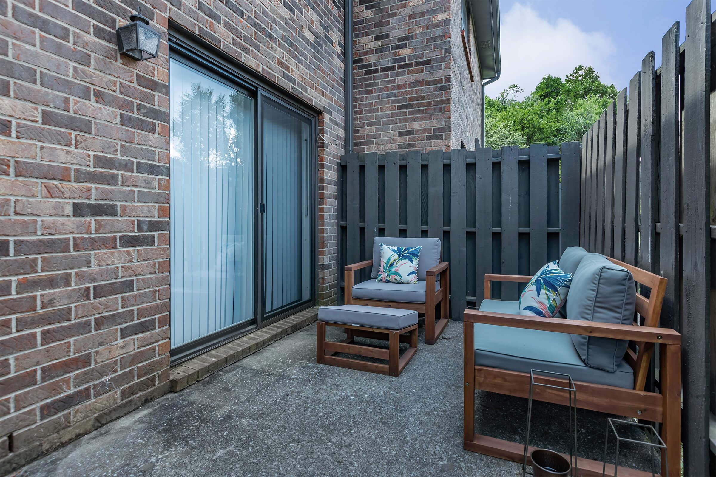 A cozy outdoor patio area featuring two wooden chairs with cushions, a small side table, and a privacy fence made of dark wood. The space has a brick wall backdrop and large sliding glass doors leading indoors, with greenery visible in the background.