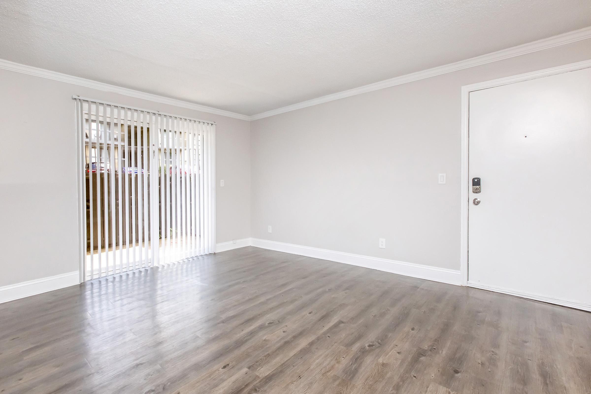 Empty room with light gray walls and a light wood floor. A white door is visible on the right, and sliding glass doors with vertical blinds are on the left, allowing natural light to enter. The space appears clean and minimalistic, suitable for furnishing.