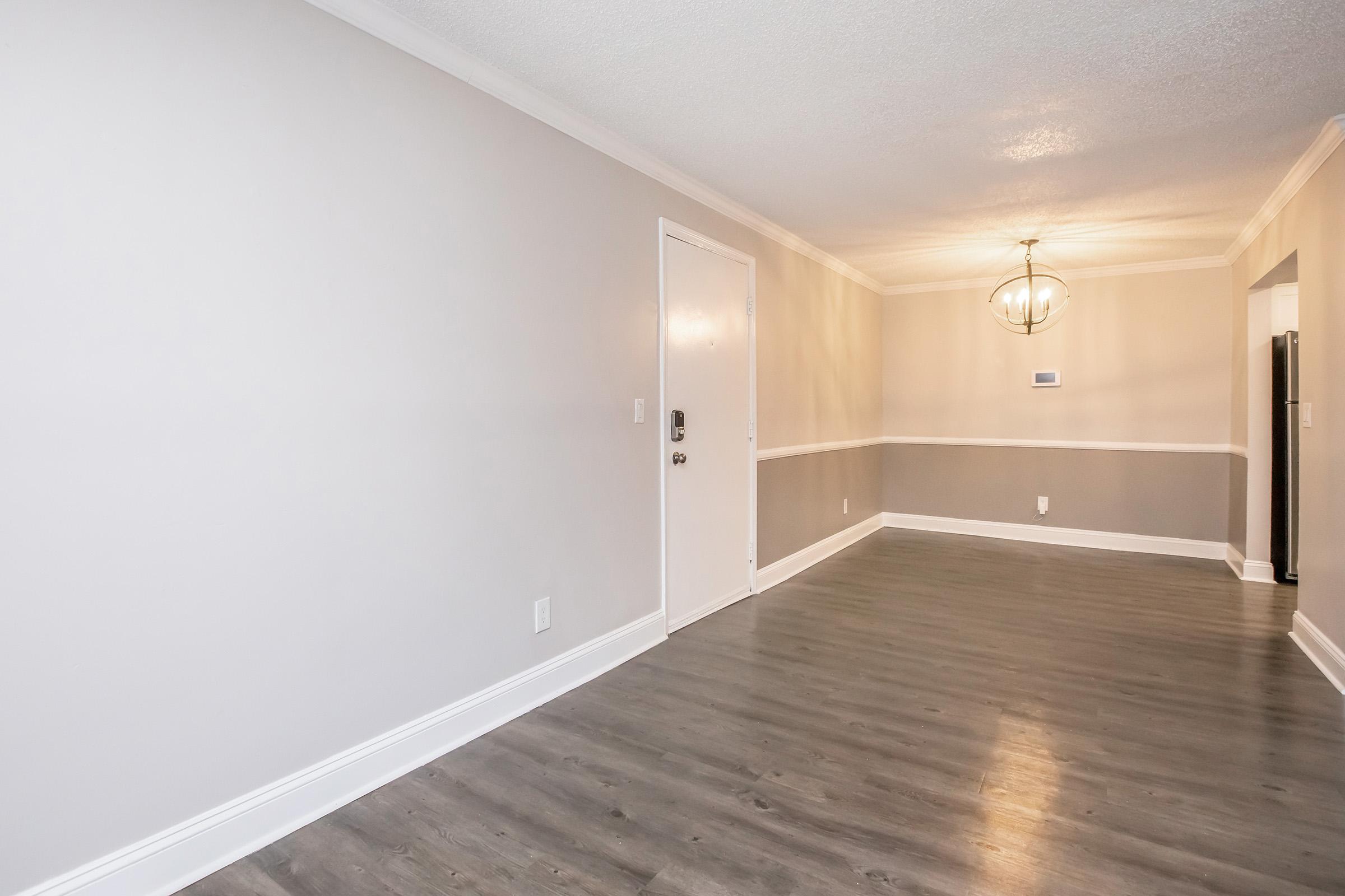 A spacious, modern living room featuring light gray walls and hardwood flooring. The entrance door is visible on the left, with a decorative light fixture hanging from the ceiling. The room is empty, providing a blank canvas for decoration and furnishings, with a subtle division created by a wall trim.