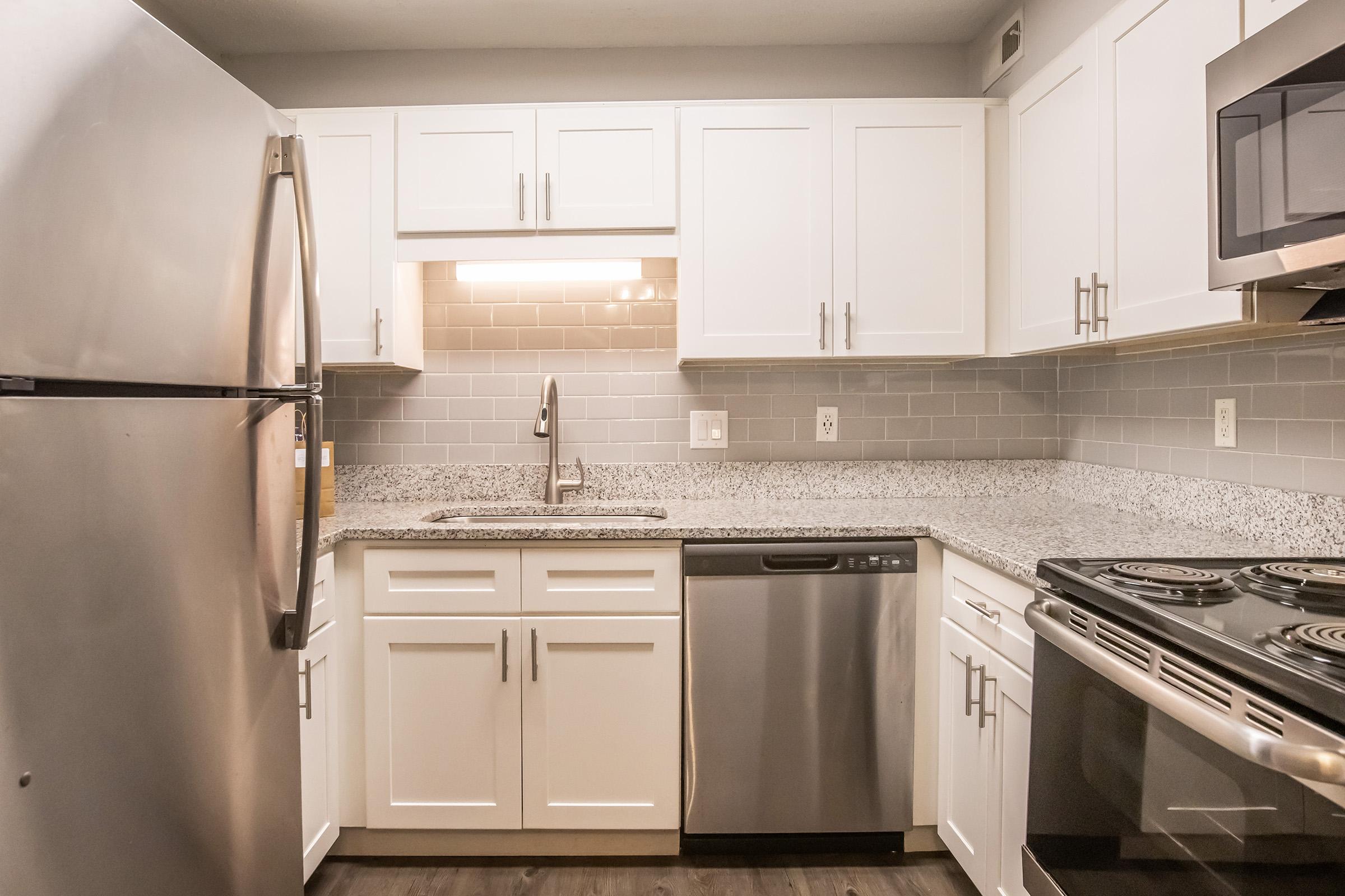 Modern kitchen featuring stainless steel appliances, including a refrigerator and oven. The white cabinetry contrasts with the granite countertops and gray tile backsplash. A sink is installed under a light fixture, providing a bright workspace. The flooring is dark laminate, complementing the overall design.