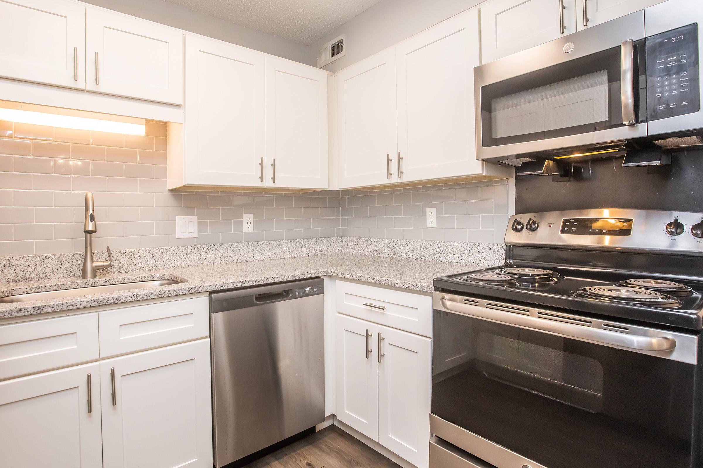 Modern kitchen featuring white cabinetry, a stainless steel dishwasher, a sleek oven with a black finish, and a microwave above it. The countertops are made of gray speckled granite, with a tile backsplash in light gray. Soft lighting enhances the clean and contemporary aesthetic.