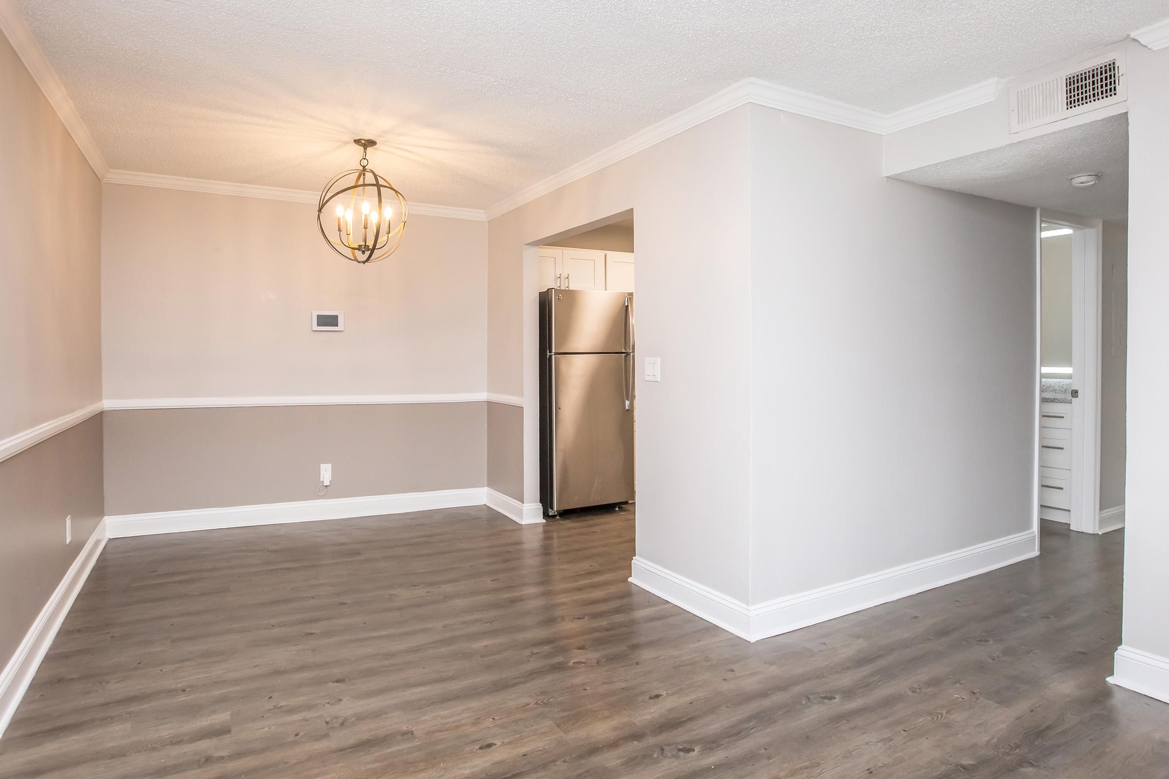 Interior view of a modern living space featuring a stylish light fixture, neutral-colored walls, and laminate flooring. A kitchen area with a stainless steel refrigerator is visible in the background, along with a doorway leading to another room. The space is well-lit and inviting.