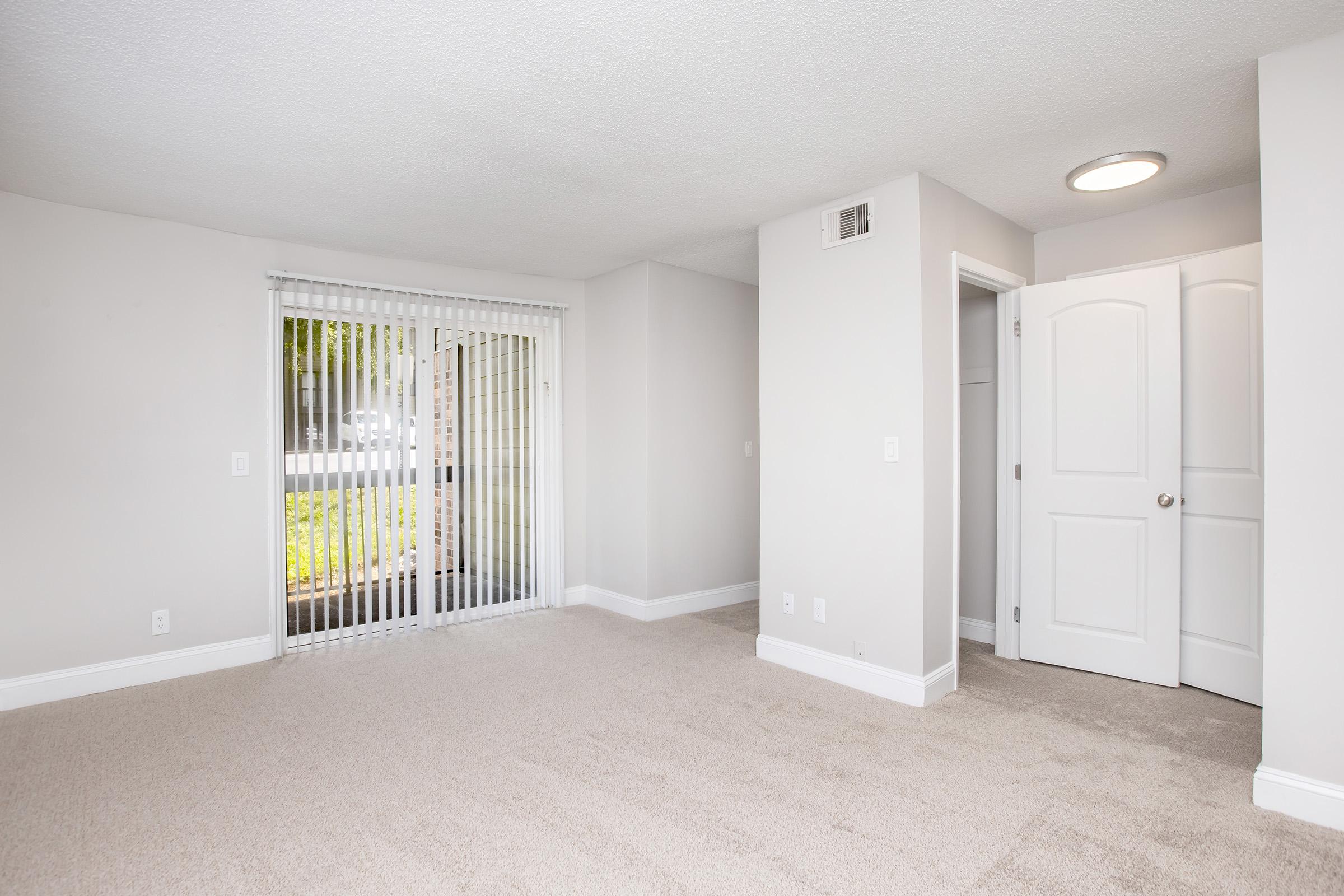 A bright, empty room with light beige carpet, light gray walls, and a sliding glass door featuring vertical blinds. A white interior door is visible on the right side, and there’s a ceiling light fixture. The large window allows natural light to enter, creating an inviting and open atmosphere.
