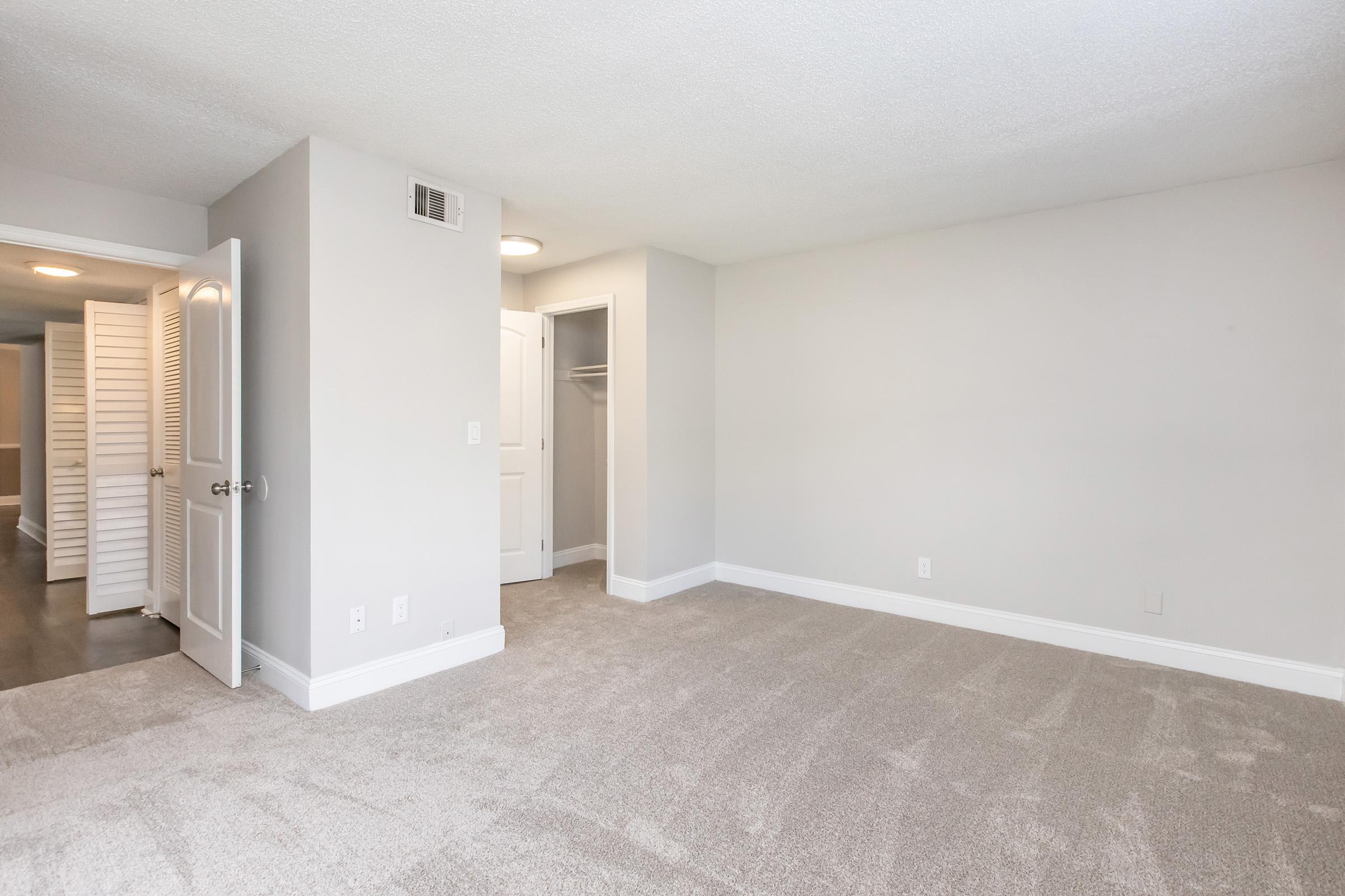 A spacious, well-lit room featuring light gray walls and plush carpeting. The room includes a closet with white louvered doors and access to another area through a closed door. The floor is clean and free of clutter, creating a tranquil and inviting atmosphere.