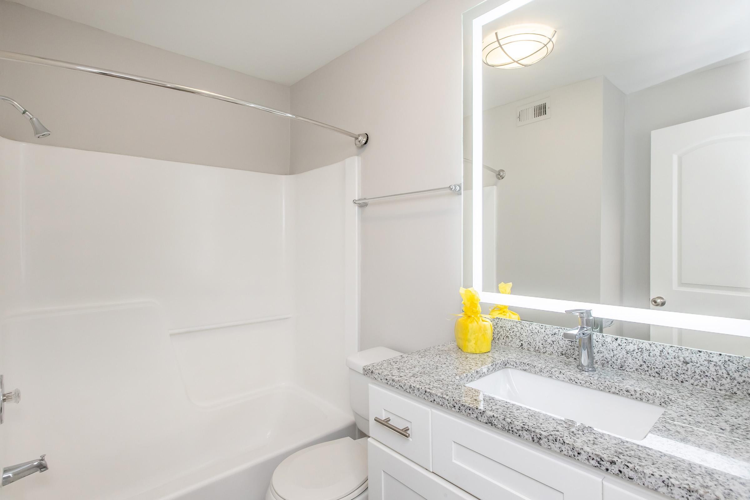A modern bathroom featuring a white bathtub with a curved shower rod, a granite countertop with a white sink, and a mirror above. Accents include a bright yellow decorative item near the sink, and a soft gray wall color. A clear reflection can be seen in the mirror, enhancing the sense of space.