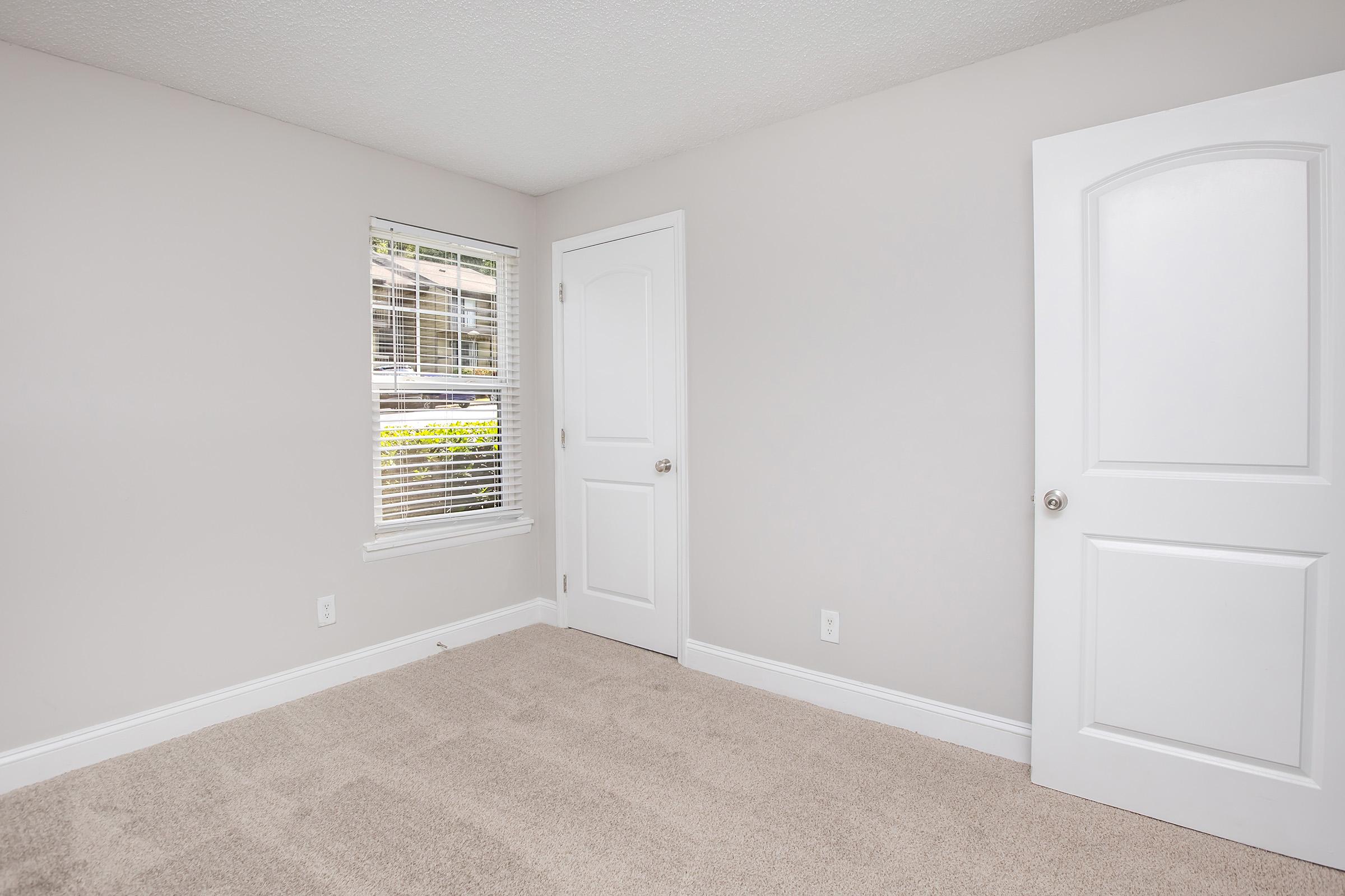 Empty room with light beige walls and a plush carpet. A window with white blinds allows natural light to enter. There are two doors: one leading to another room and the other possibly a closet. The overall space is bright and uncluttered, creating a calm atmosphere.