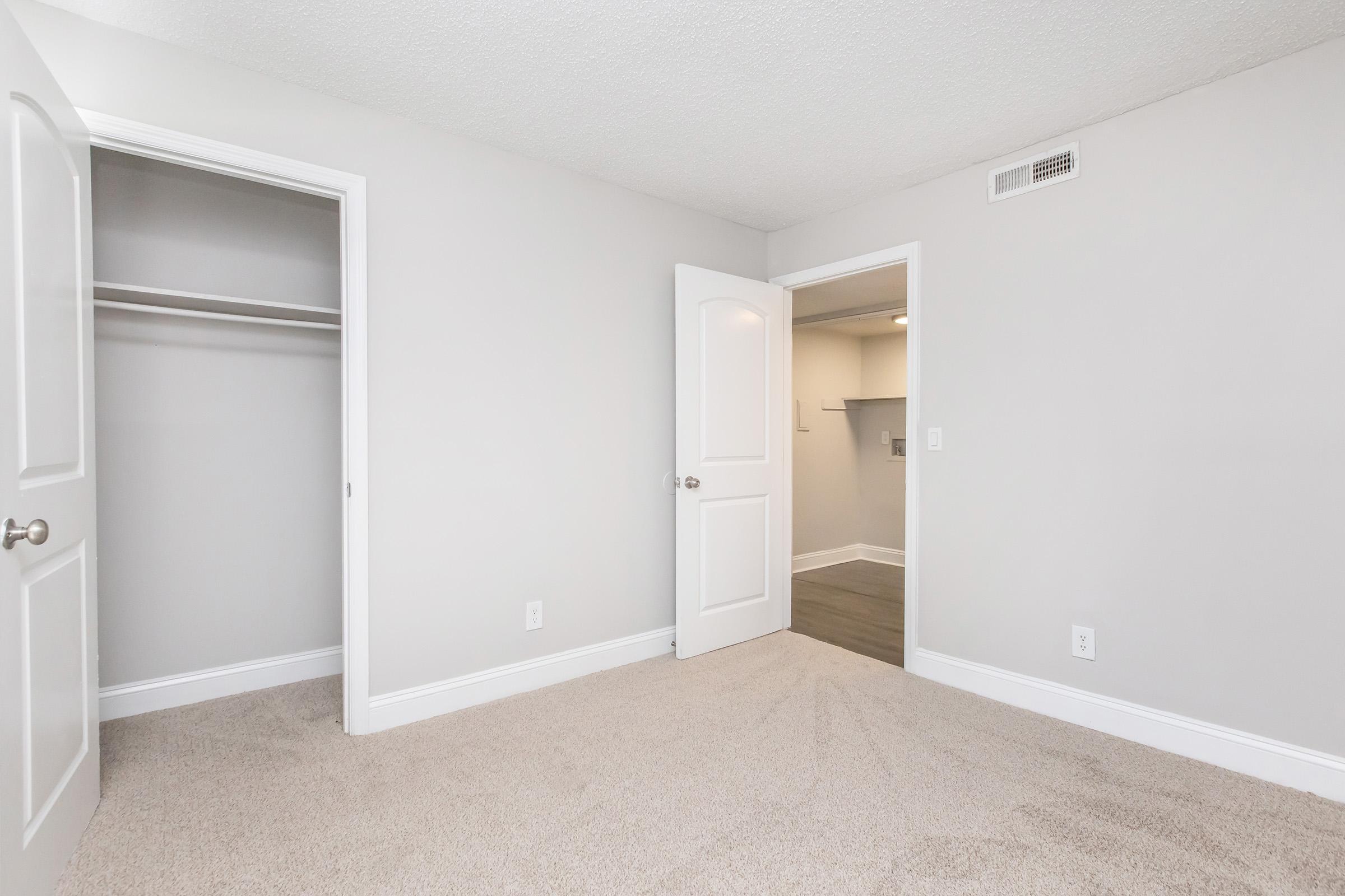 A light gray room with two open white doors. One door leads to a closet with no visible contents, while the other opens to another room with a partially visible wall. The carpet is beige, and the walls are painted in a neutral tone. The lighting creates a bright and airy atmosphere.