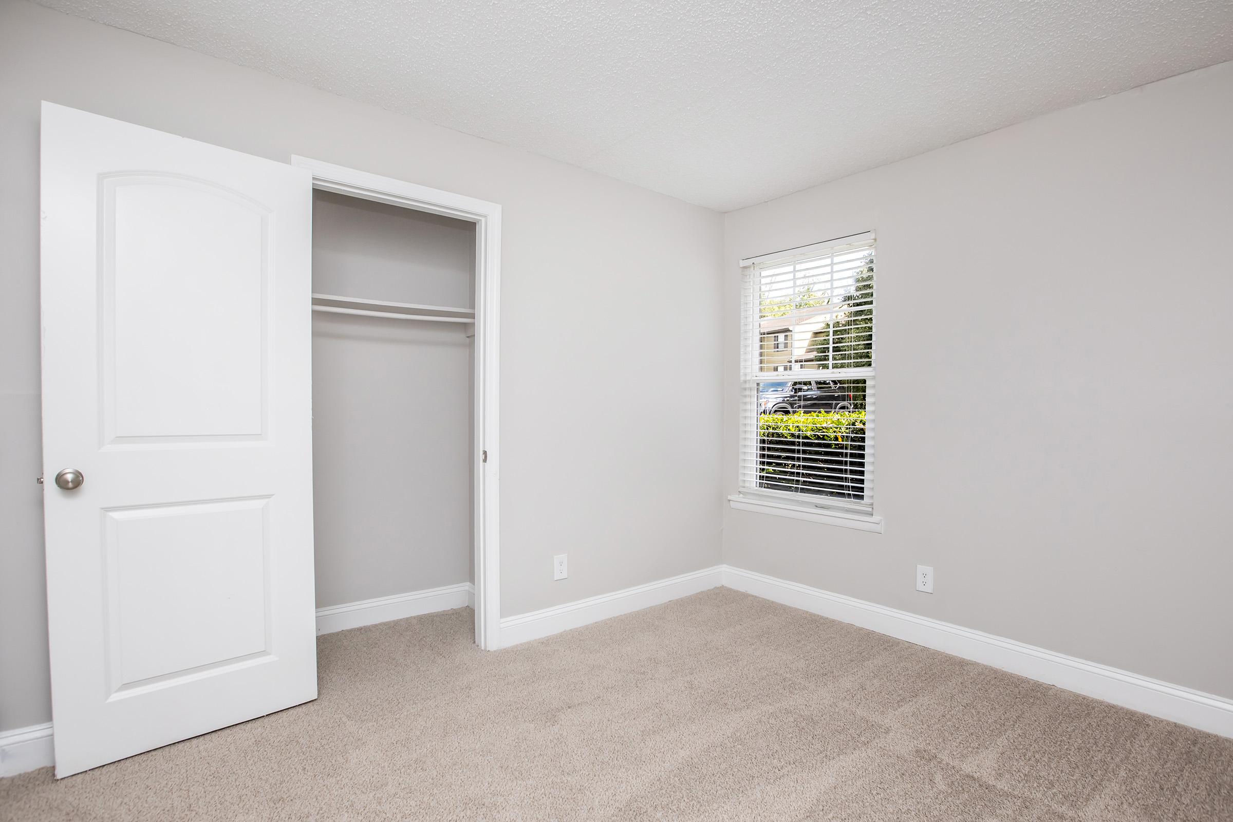 A quiet bedroom with light gray walls and beige carpet. The room features a closed white door leading to a closet, a window with white blinds allowing natural light in, and a view of greenery outside. The space is empty and bright, creating a feeling of openness.
