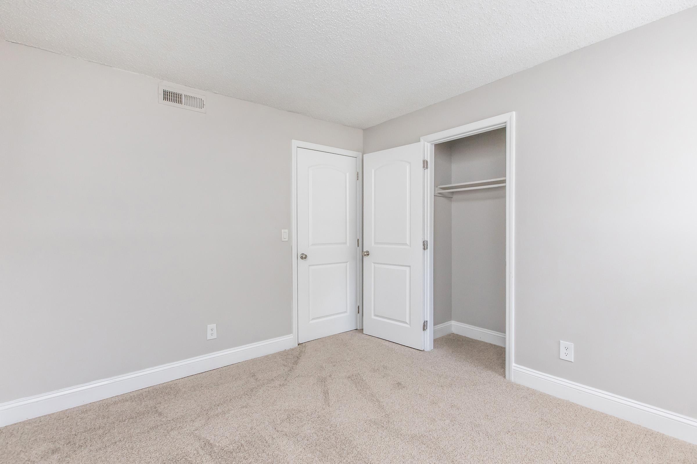 A neatly maintained bedroom featuring light gray walls and a plush carpet. There are two white doors: one leads to a closet, and the other opens into an adjacent room. The space is bright and airy, with no furniture present, creating an open, inviting atmosphere.