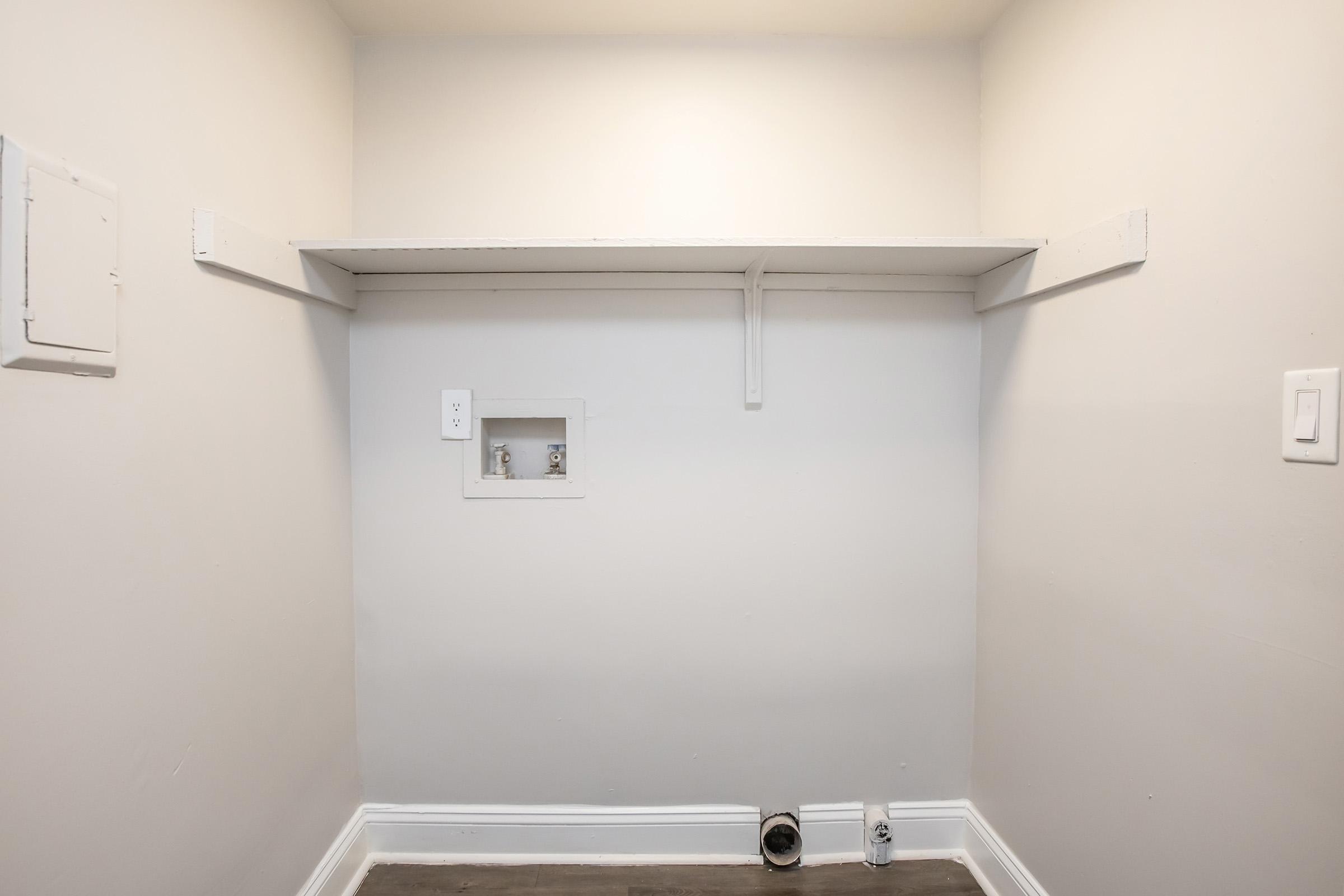 Empty wall with a white shelf supported by brackets. A small recessed space is visible, possibly for storage or utilities, and there are two electrical outlets on either side of the shelf. The room is painted in a light color, contributing to a clean and minimalistic appearance.