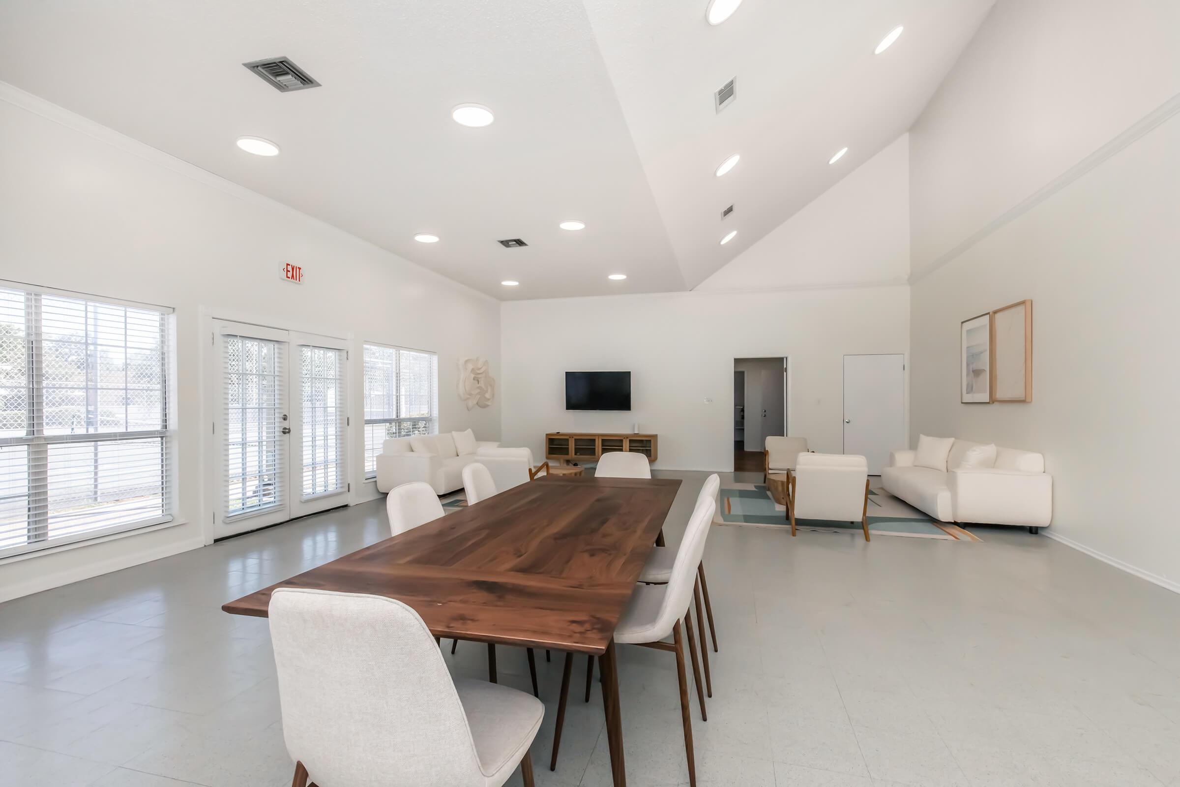 A modern, spacious living area featuring a large wooden dining table with white chairs, a comfortable seating area with light-colored sofas, and a wall-mounted television. Large windows allow natural light, creating a bright atmosphere in the neutral-toned room.