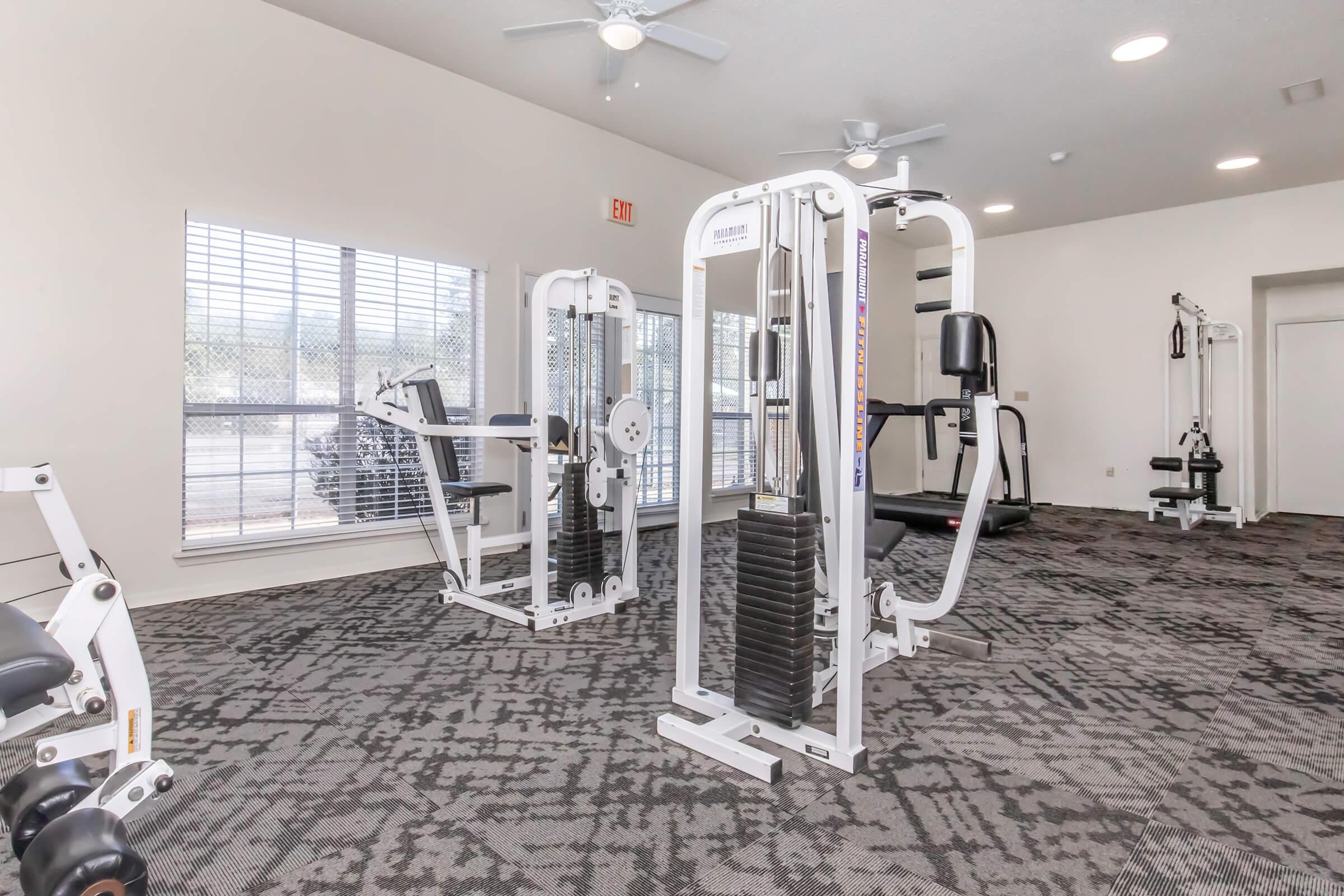A well-lit gym interior featuring multiple exercise machines, including a leg press and weight stack machines. The room has soft, neutral-colored walls and large windows allowing natural light, with a modern carpet design below. Ceiling fans are installed for ventilation.