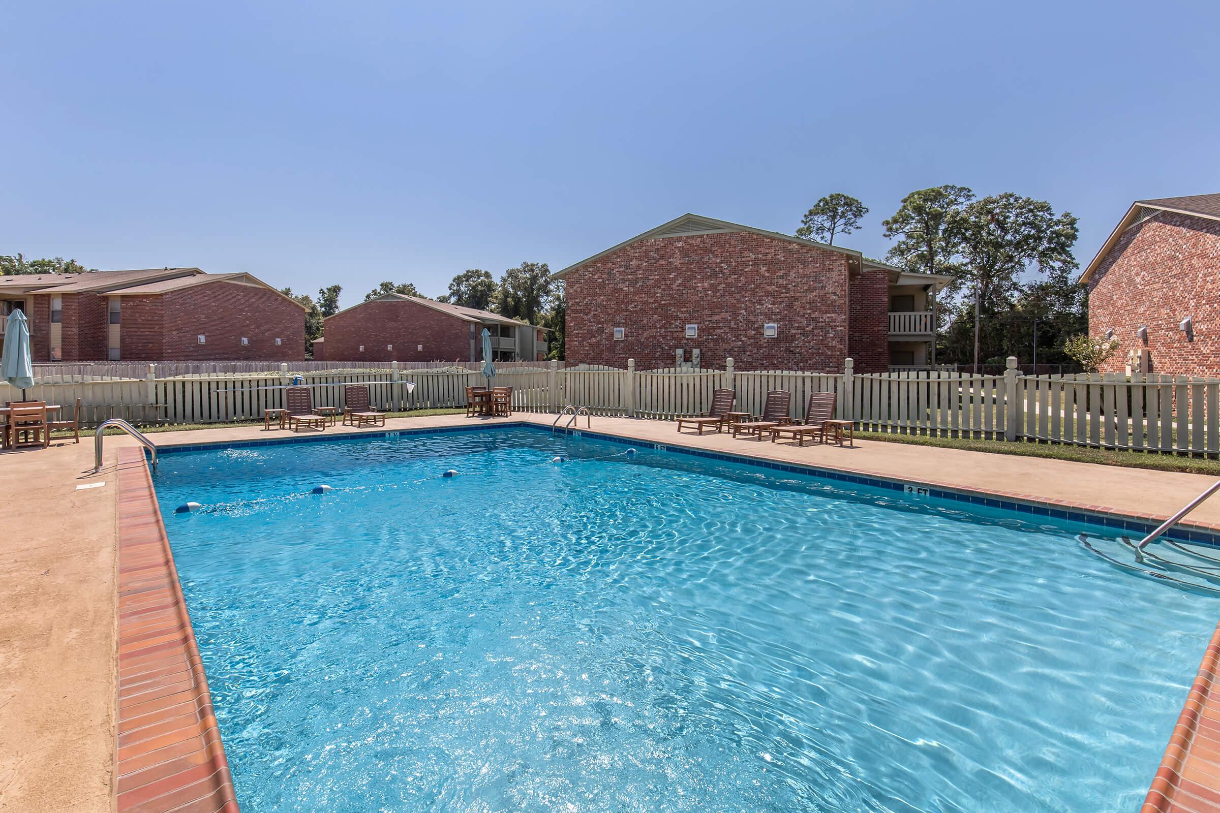 A bright blue swimming pool surrounded by a fenced area, with lounge chairs on the deck. In the background, two brick apartment buildings are visible under a clear sky. The setting is inviting and spacious, perfect for relaxation and leisure activities.