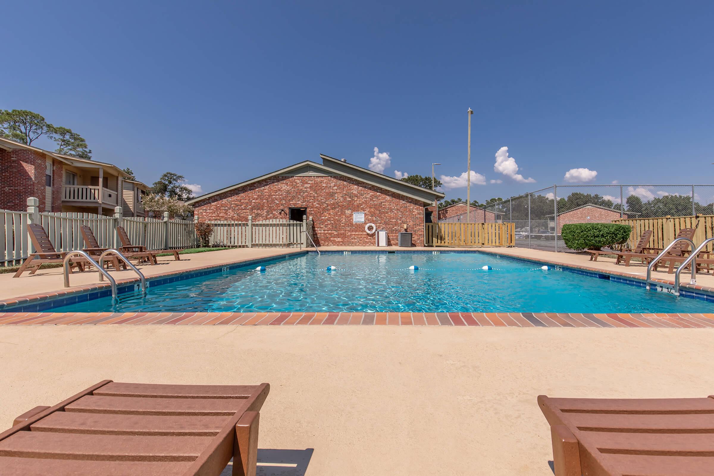 A clean, inviting outdoor swimming pool surrounded by lounge chairs, with a wooden fence in the background. There are two buildings visible, set against a clear blue sky with a few clouds. The pool area includes a swimming lane marked by buoys.