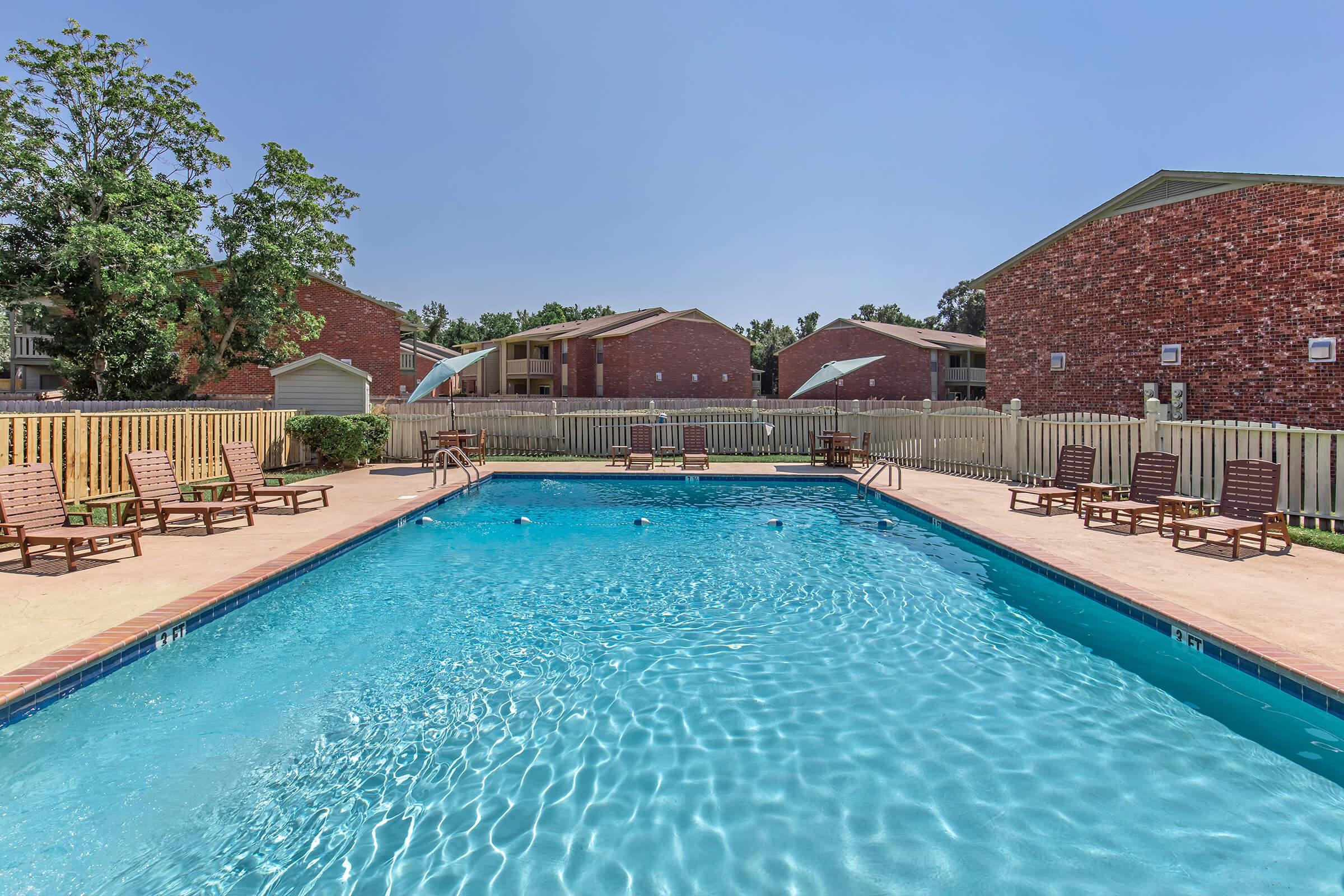 A clear blue swimming pool surrounded by lounge chairs, with green trees and brick apartment buildings in the background. The area is enclosed by a wooden fence, and the sky is bright and sunny, creating a relaxing outdoor atmosphere.