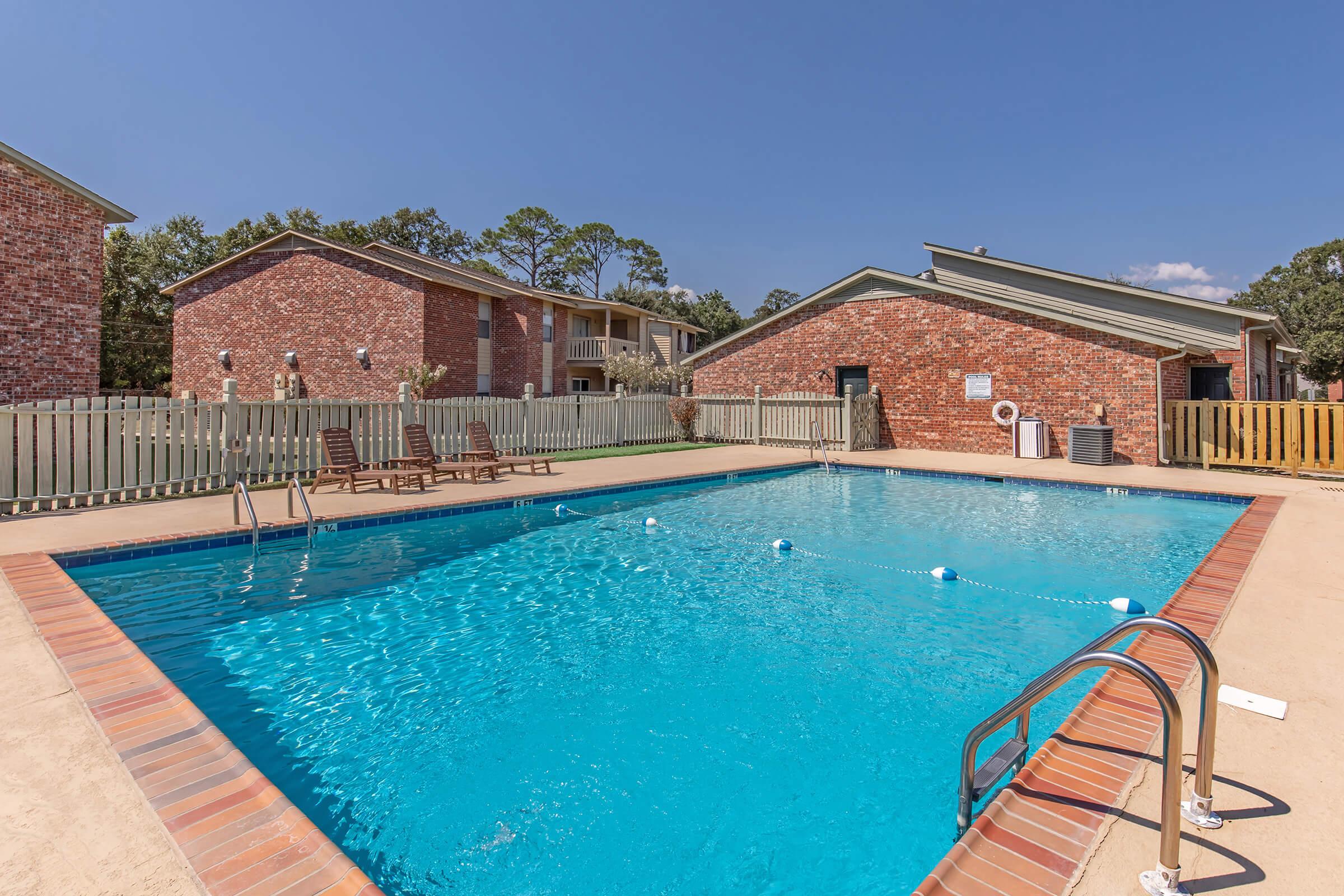A sunny outdoor swimming pool surrounded by a brick-paved area. There are several lounge chairs positioned nearby, with wooden fencing and residential buildings in the background. Clear blue water fills the pool, and the sky is bright and blue, creating a relaxing atmosphere.