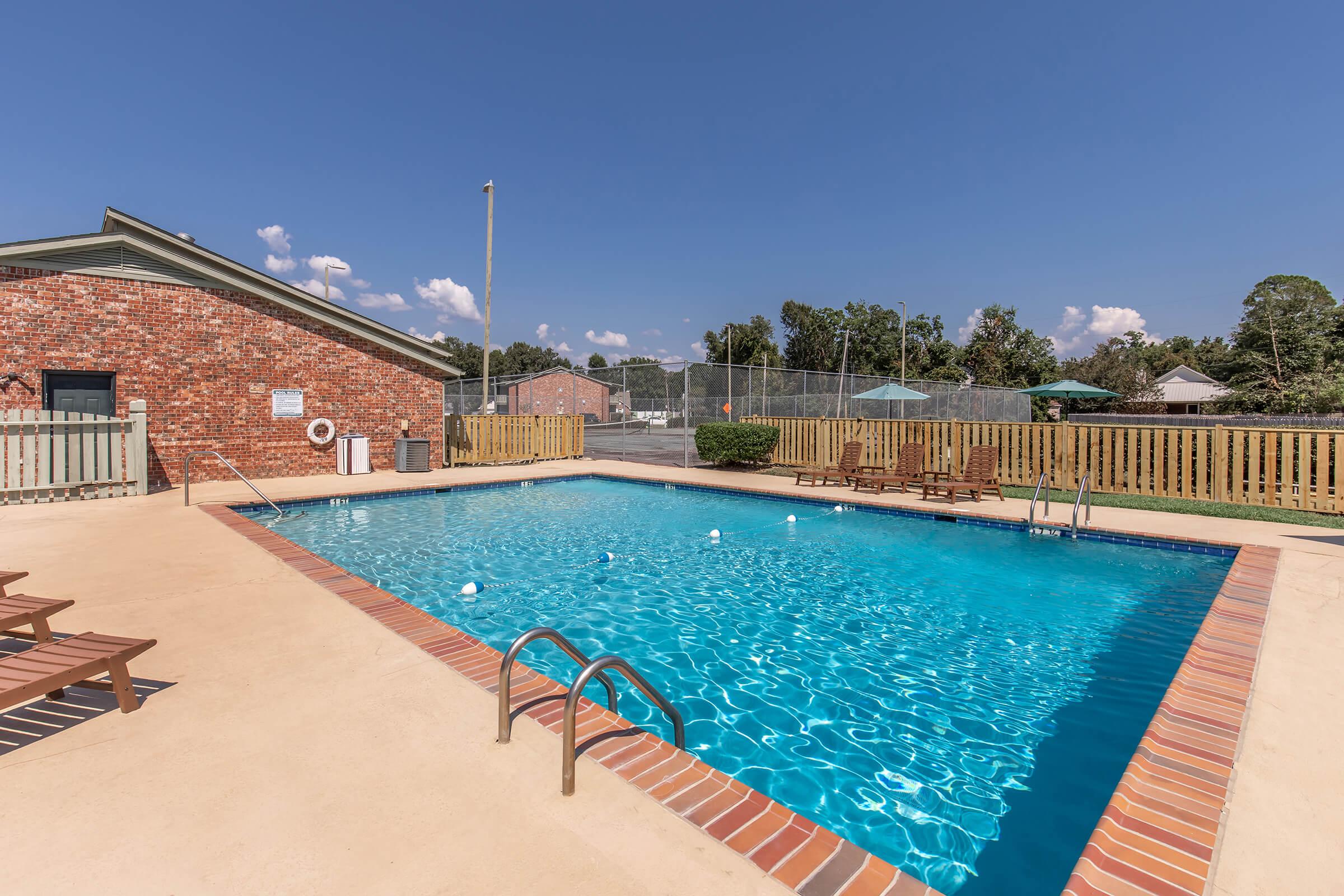 A clear swimming pool with bright blue water surrounded by a deck, lounge chairs, and a fence. In the background, a tennis court is visible. The area is sunny with a few clouds in the sky, and a building is seen nearby. Ideal for relaxation and leisure activities.