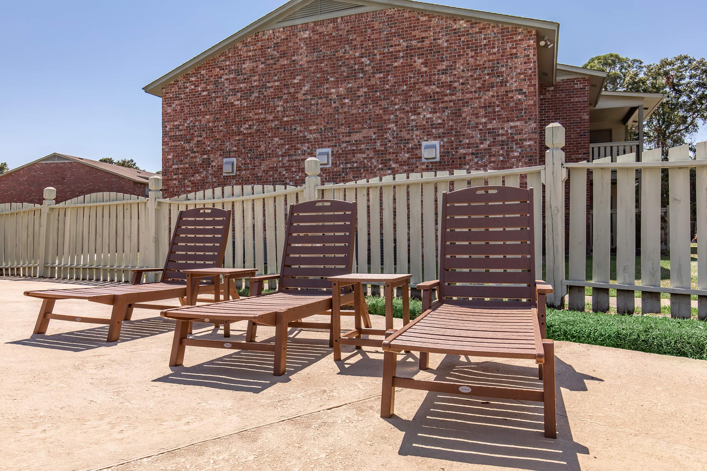 Four wooden lounge chairs are positioned on a patio in front of a fenced area, with a brick building visible in the background. The scene is sunny, and the chairs are arranged neatly, suggesting a relaxing outdoor space.