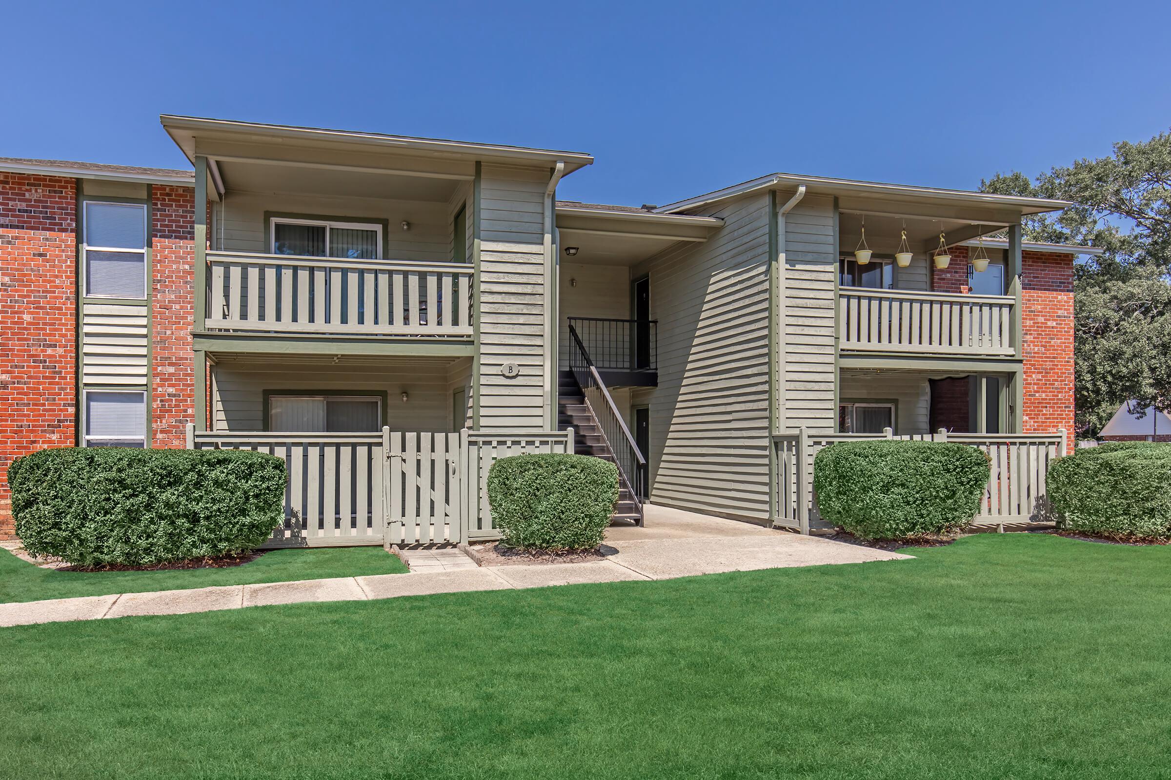 Two-story apartment building with a light gray exterior, featuring balconies and a central staircase. The building is surrounded by well-maintained green grass and small hedges, under a clear blue sky.