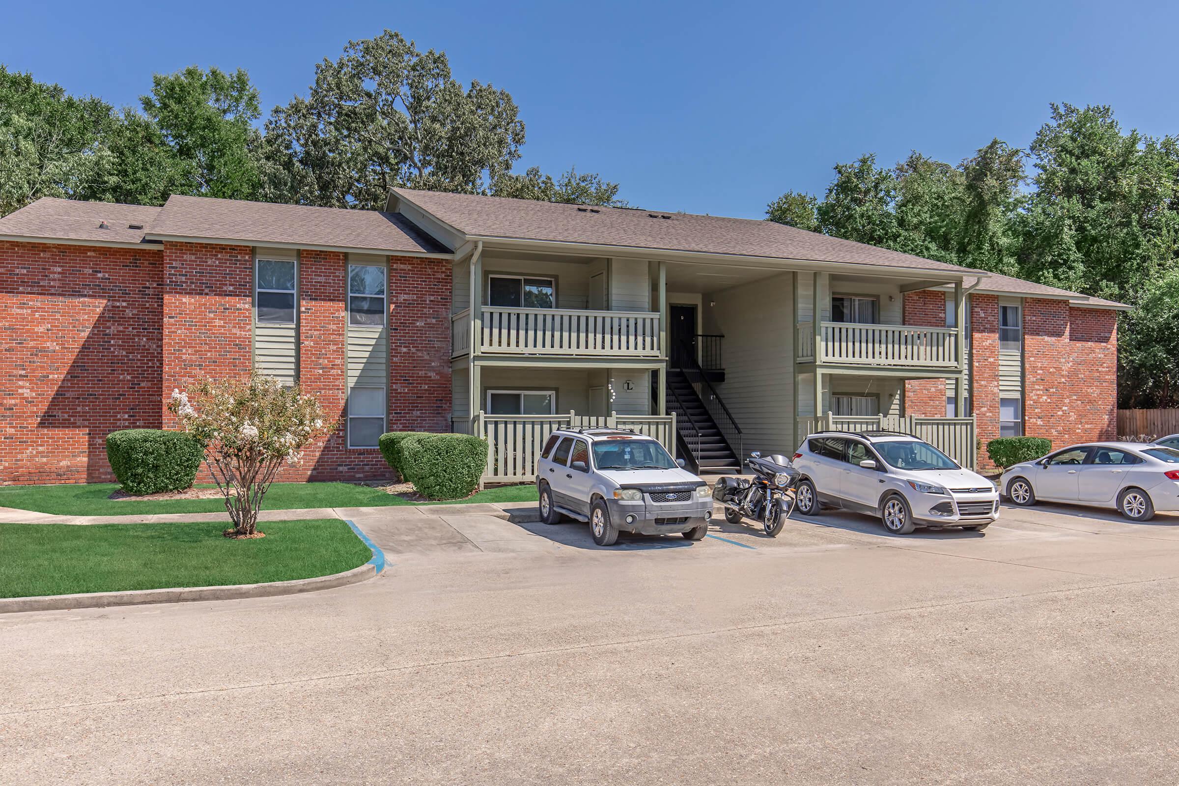 A brick apartment building with multiple units, featuring balconies and a staircase. The parking lot has several vehicles, including cars and a motorcycle. Surrounding the building are well-maintained green lawns and trees, under a clear blue sky.