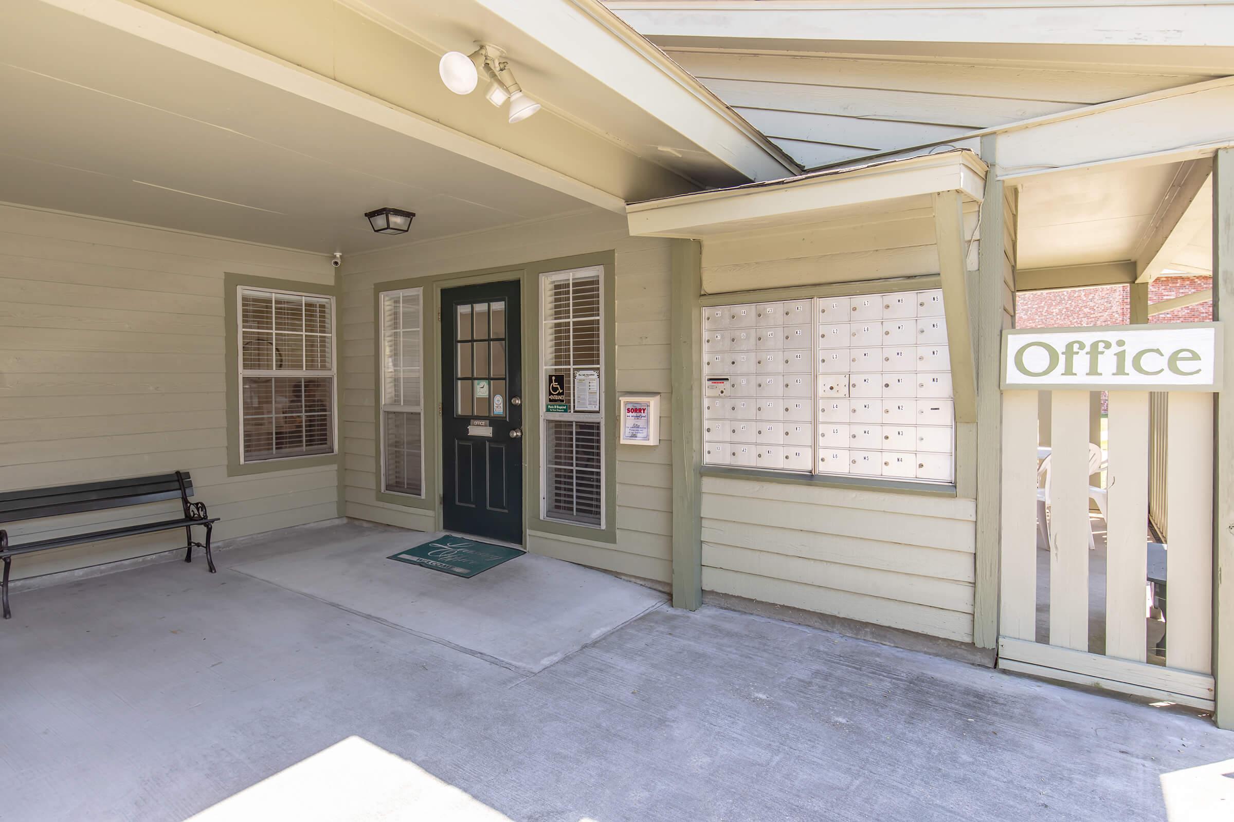 Entrance to an office building featuring a green door, window with blinds, and a row of mailboxes. There's a bench to the left and a sign indicating the location of the office. The exterior is painted in light green and tan colors, creating a welcoming atmosphere.