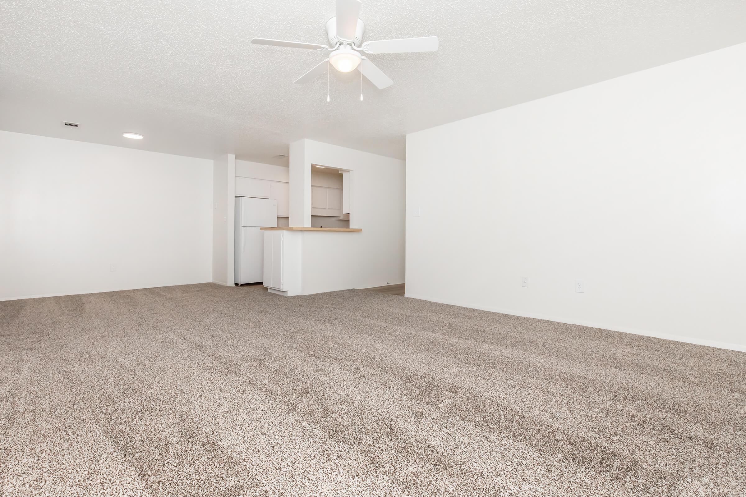 A spacious, empty living area with light-colored carpeting and a ceiling fan. The walls are painted white, and there’s a small kitchen area with a bar counter visible in the background. A white refrigerator is also seen, enhancing the bright and airy feel of the room.