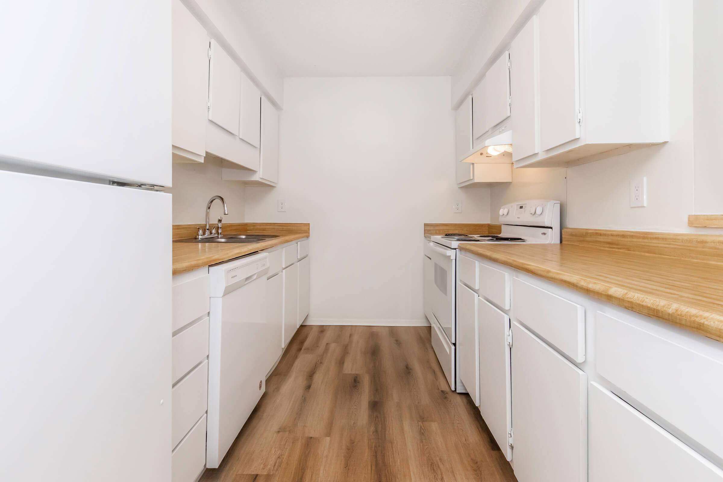 A bright and modern kitchen featuring white cabinetry and appliances, wooden countertops, and a clean, uncluttered look. The space includes a refrigerator, dishwasher, sink, and stove, with ample cabinetry for storage and a warm wood floor, creating an inviting atmosphere.