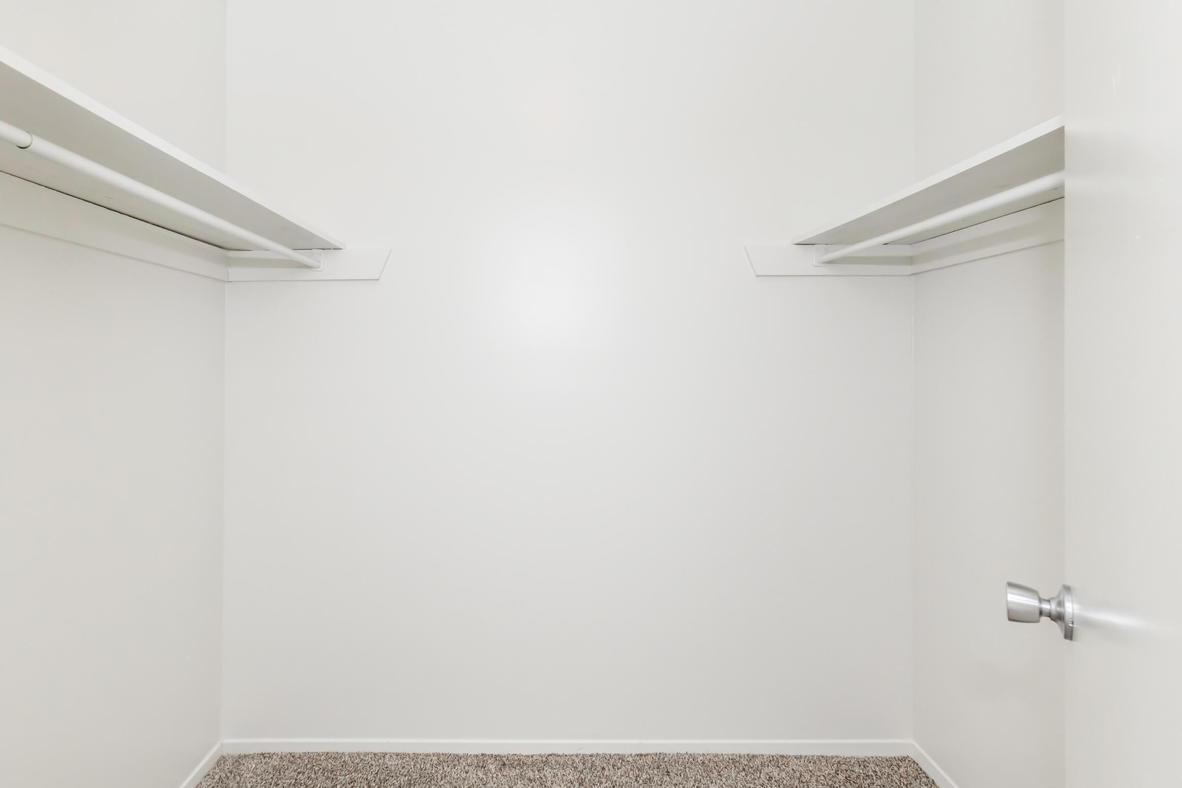 A spacious, empty closet with white walls and two shelves on either side. The floor is covered in light-colored carpet, and there's a handle on the closed door, indicating a simple, minimalist design. The overall feel is clean and uncluttered, ready for storage.