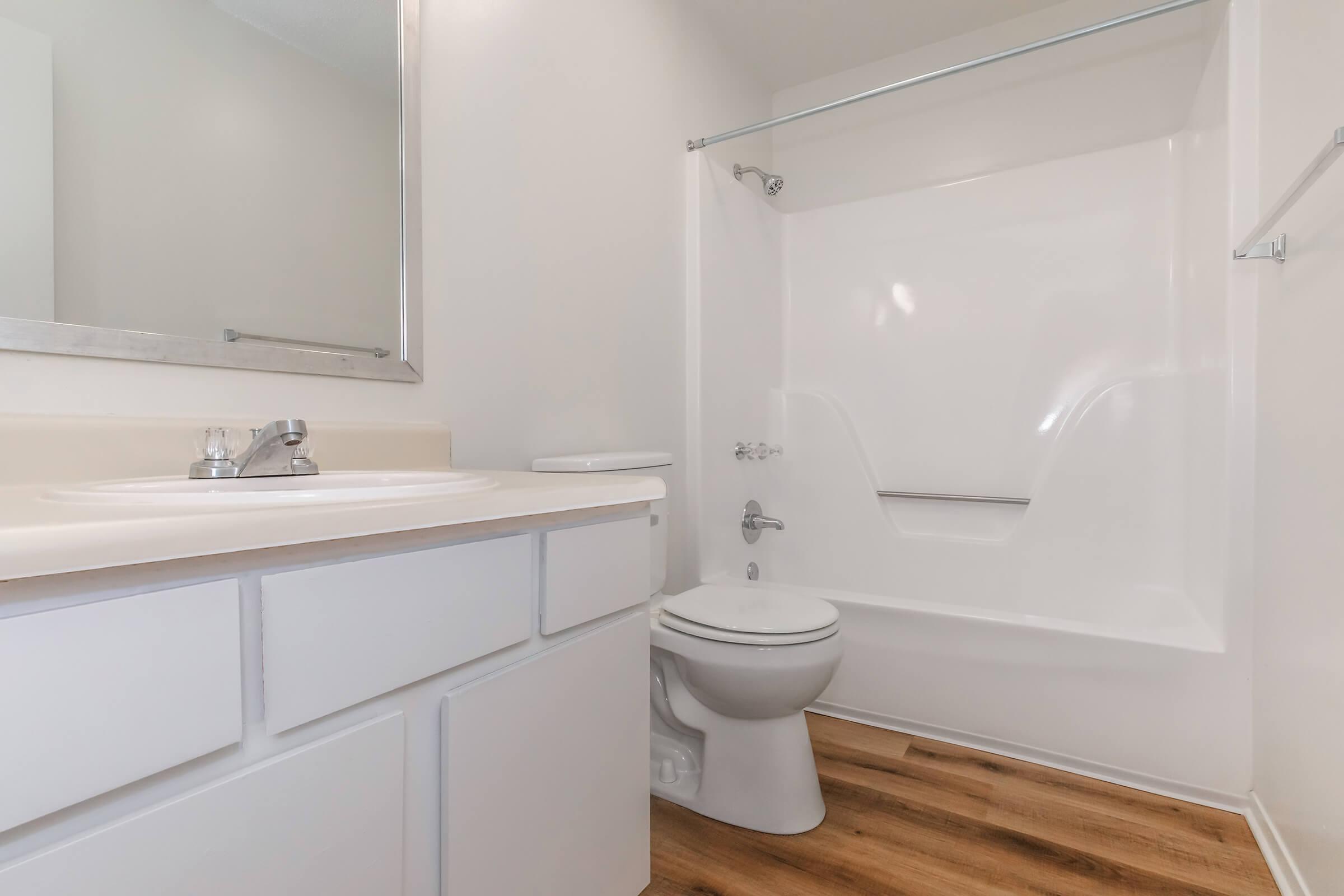 A clean and modern bathroom featuring a white bathtub with a shower, a white toilet, and a countertop with a sink. The walls are painted light gray, and there is a large mirror above the sink. The floor is made of wood-like laminate, enhancing the room's contemporary feel.