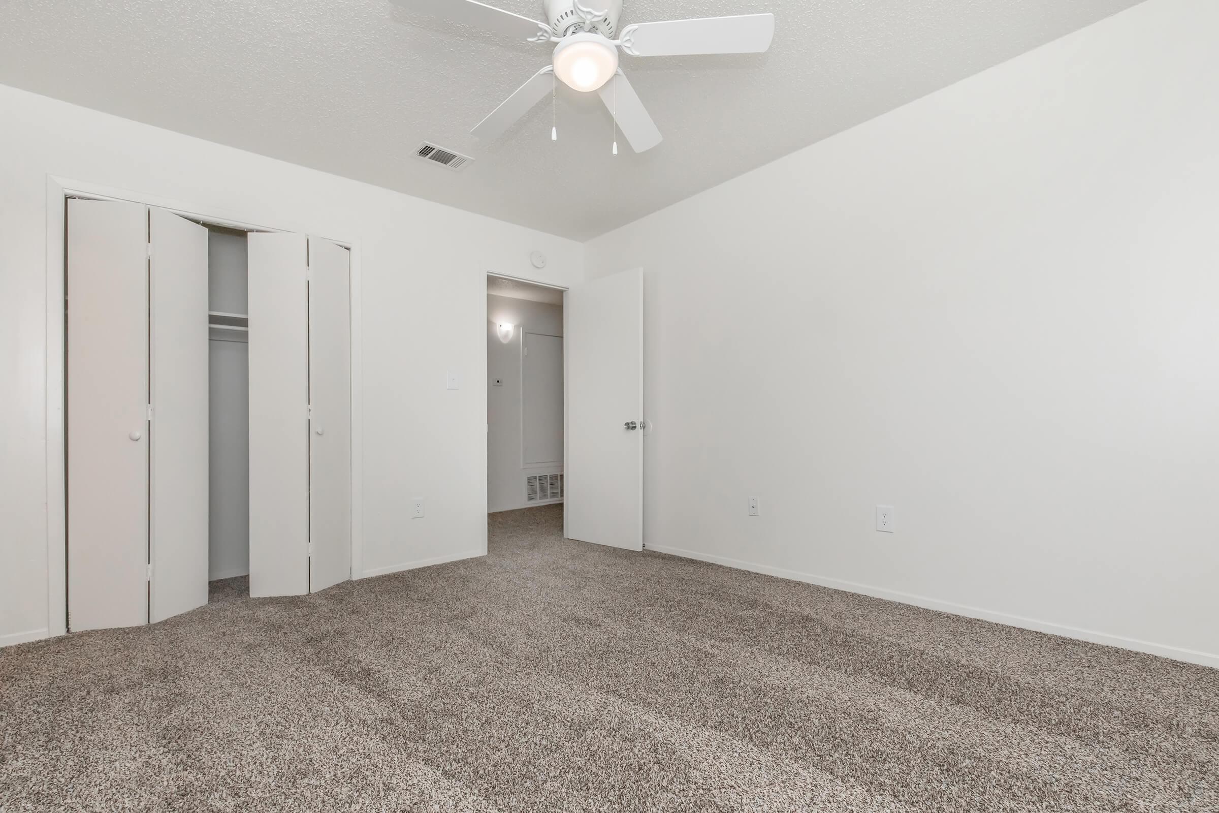 A spacious, empty room with light beige walls and a light-colored carpet. A ceiling fan is mounted above, and there are two white closet doors on the left. A door on the right leads to another room, visible in the background. Natural light brightens the space.