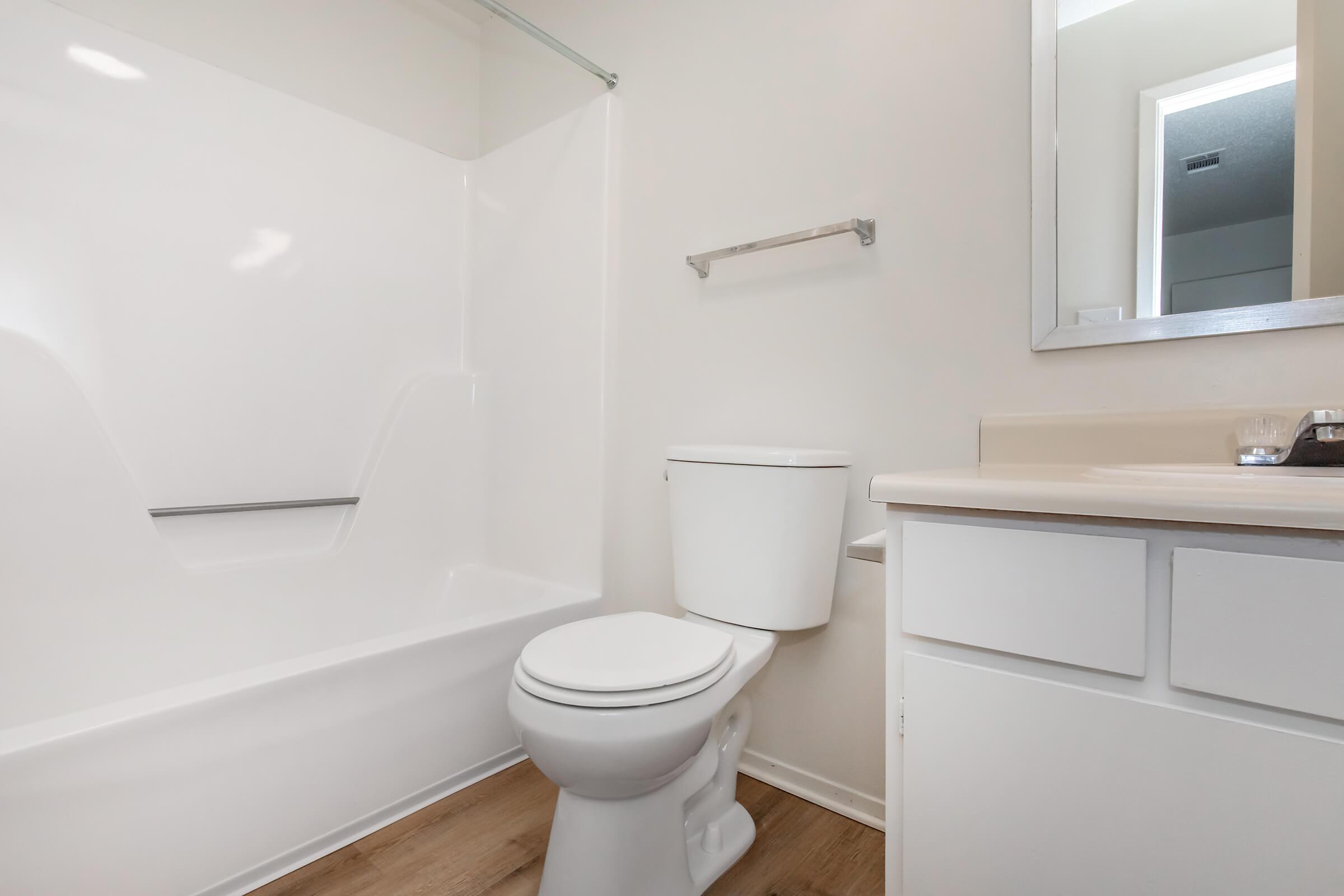 A clean, modern bathroom featuring a white bathtub with a glass shower door, a white toilet, and a countertop with a sink. The walls are light-colored, and there is a mirror above the sink, creating a bright and spacious atmosphere.