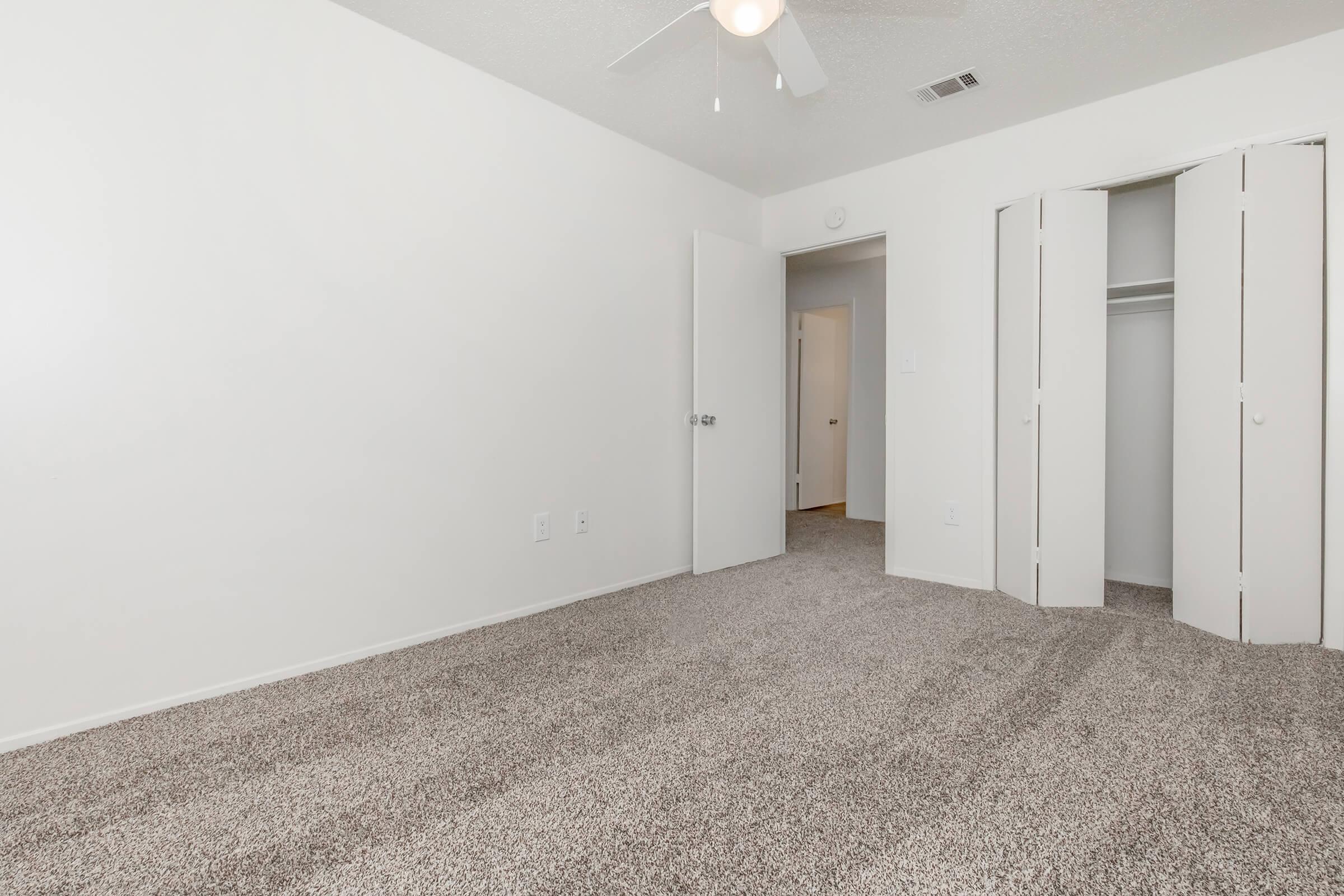 A spacious, empty bedroom featuring light-colored walls and a soft carpeted floor. A ceiling fan is visible, along with two doors: one leading to a closet and another to a hallway. The room is well-lit, creating a bright and airy feel.