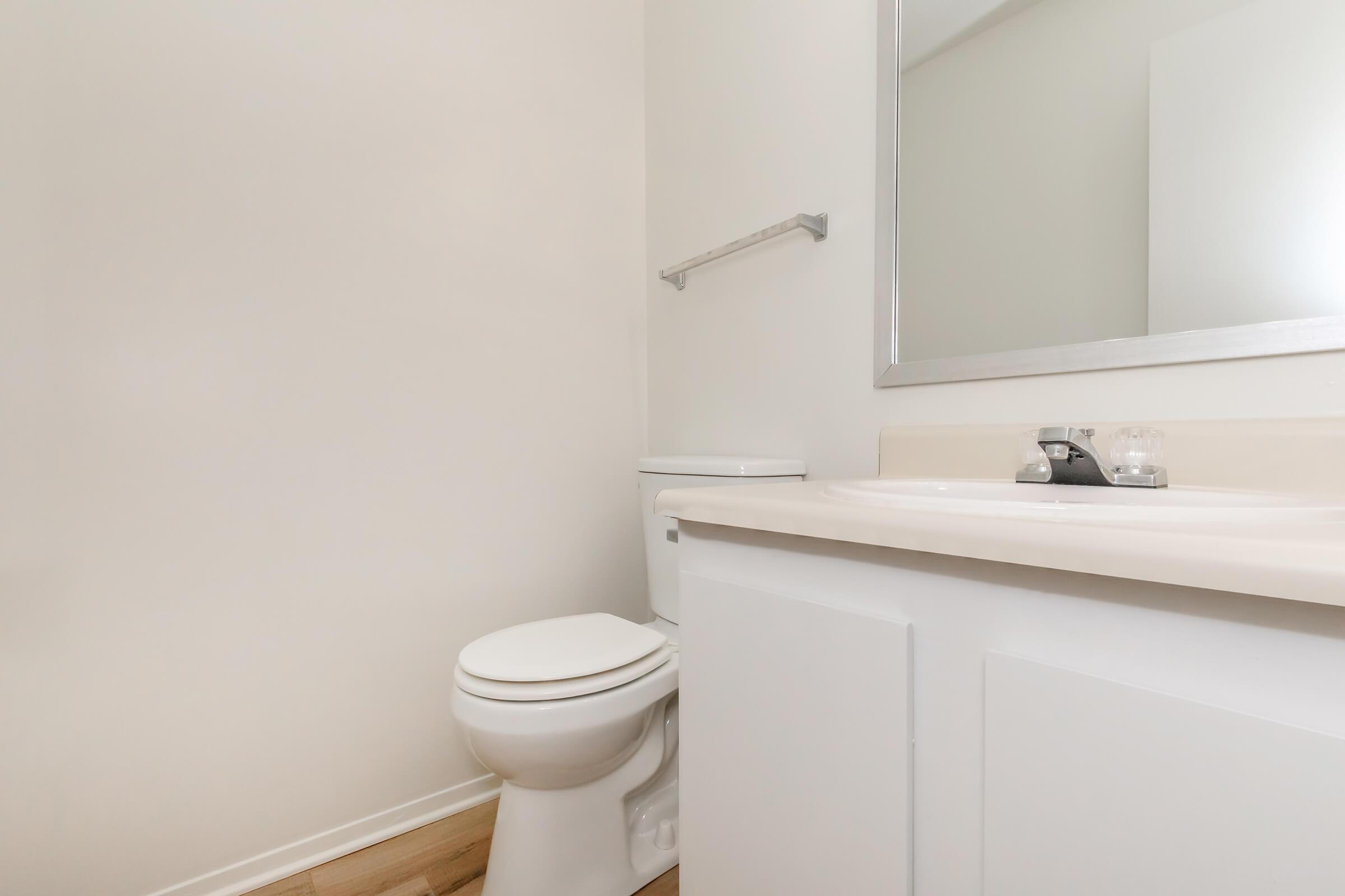 A clean, minimalist bathroom featuring a white toilet, a simple countertop with a sink, and a large mirror. The walls are painted a light color, and the flooring is wooden. The space appears bright and uncluttered, focusing on functionality.