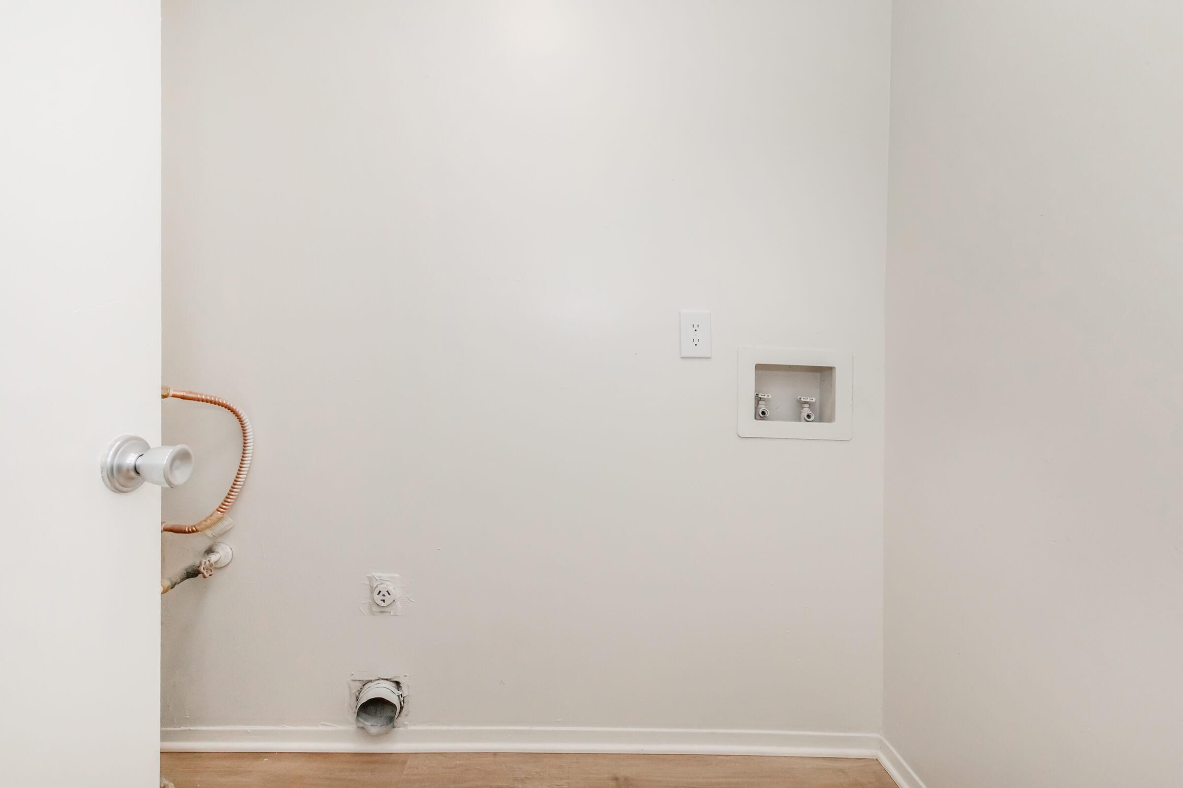 Empty interior wall with a light switch, electrical outlet, and plumbing connections visible. There is a white door on the left side and a vent at the floor level. The room has a neutral color scheme with light wood flooring.