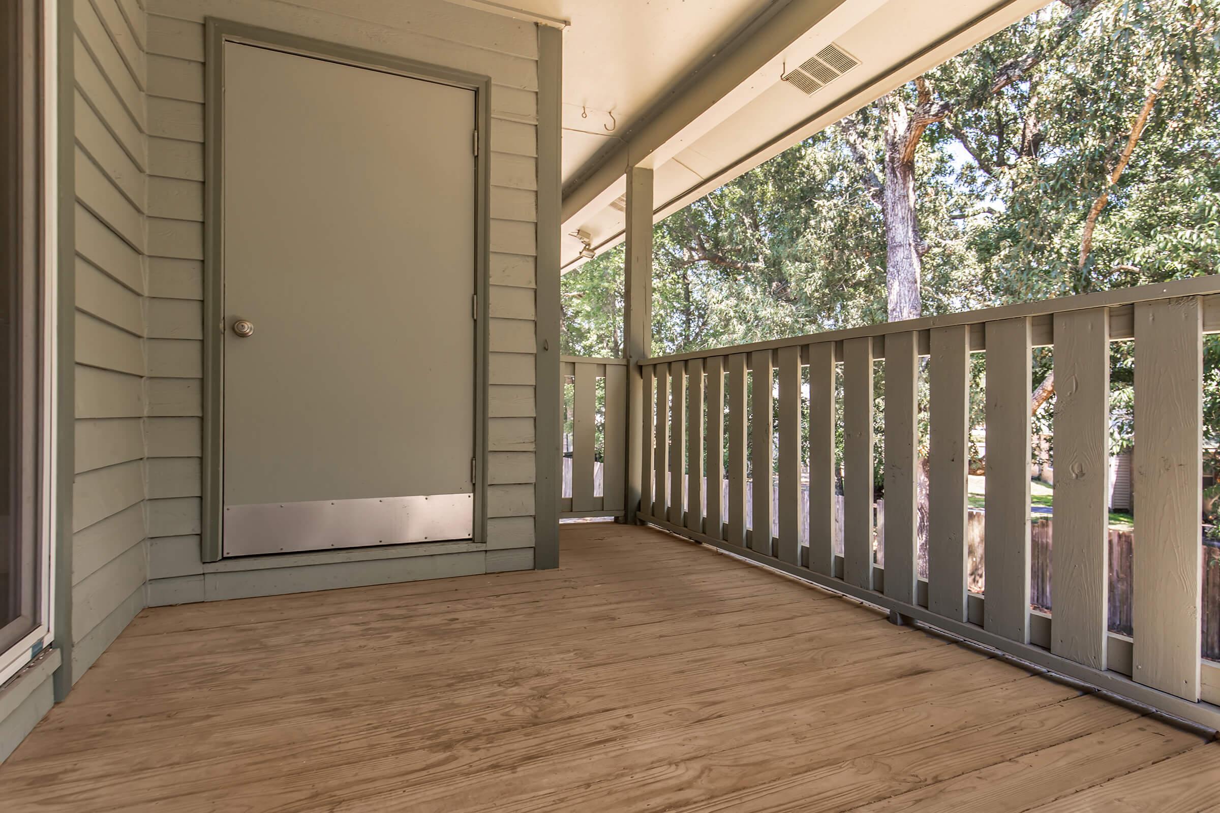A wooden porch with a light green door and white railing, surrounded by trees. The floor is made of wood, providing a natural, inviting look. The scene is well-lit, suggesting a peaceful outdoor area.