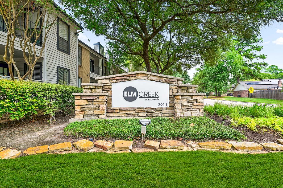 Sign for Elm Creek Apartments, featuring the name prominently displayed against a stone background. Surrounding the sign are well-maintained shrubs and a grassy area, with trees providing shade in the background. A paved road is visible in the distance.