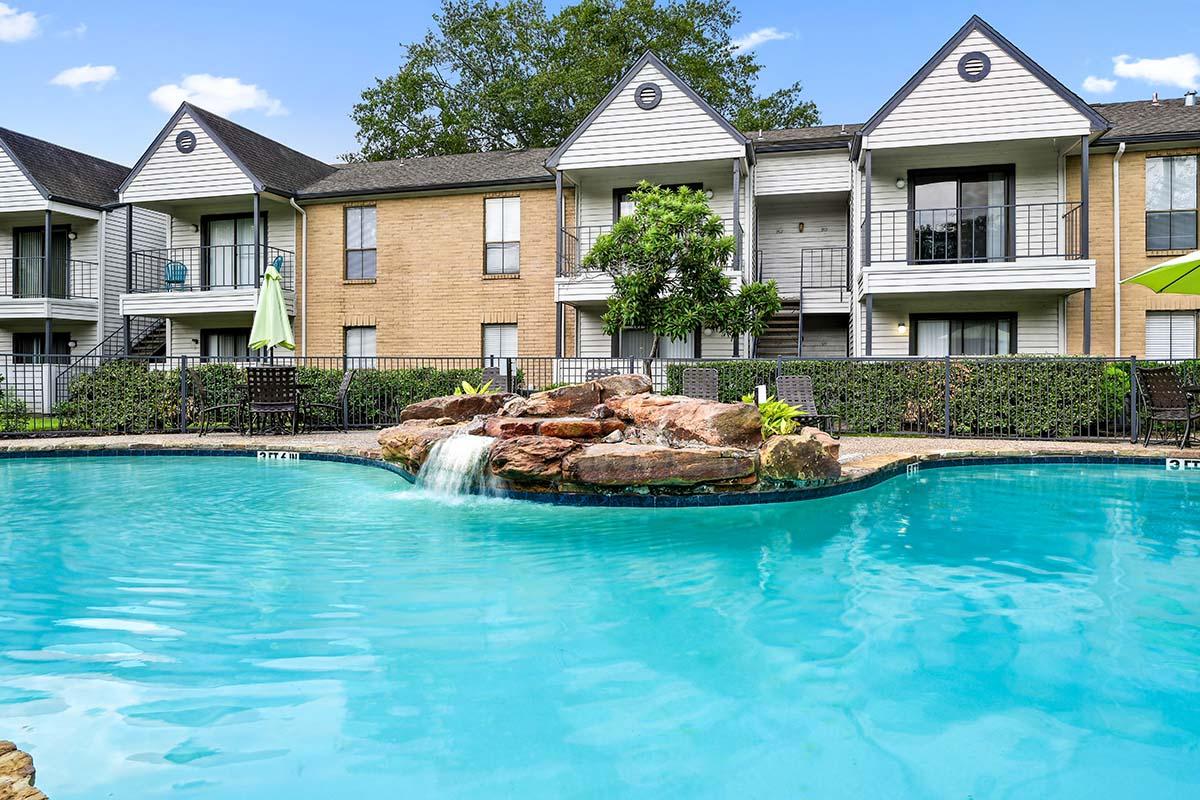 A clear blue swimming pool with a rocky fountain feature in the foreground, surrounded by lush landscaping. In the background, there are two apartment buildings with balconies and green umbrellas near the pool area, under a bright blue sky.