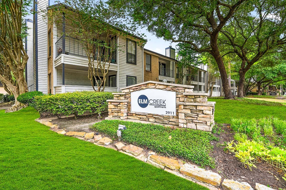 Sign for Elm Creek Apartments, featuring a stone base and white background, situated in front of well-maintained greenery and landscaping. In the background, there are two residential buildings with balconies, surrounded by trees and lush grass.
