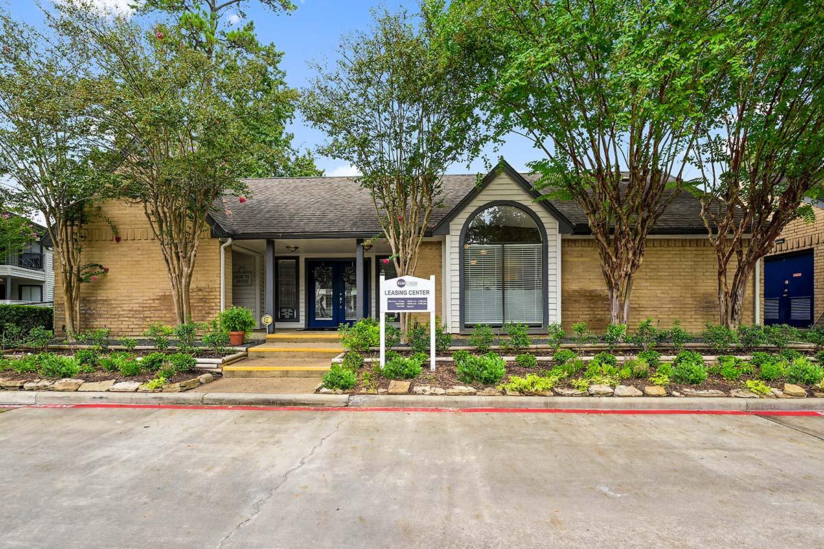 A single-story building with a sloped roof, surrounded by trees and manicured landscaping. In front, there's a sign indicating a leasing center. The entrance features steps leading up to the door, and the overall atmosphere is welcoming and well-maintained.