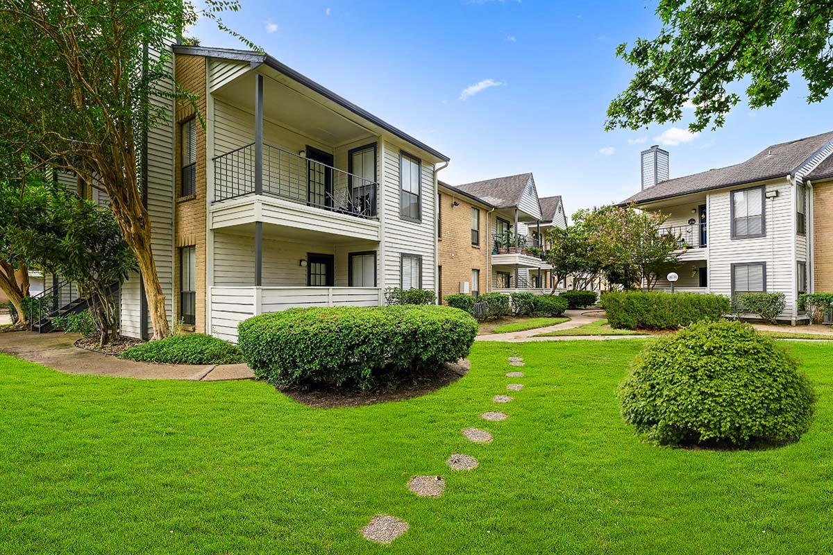 A view of a well-kept apartment complex with two-story buildings, surrounded by lush green lawns and neatly trimmed bushes. A stone pathway winds through the grass, leading to the entrances of the apartments, under a clear blue sky with a few clouds.