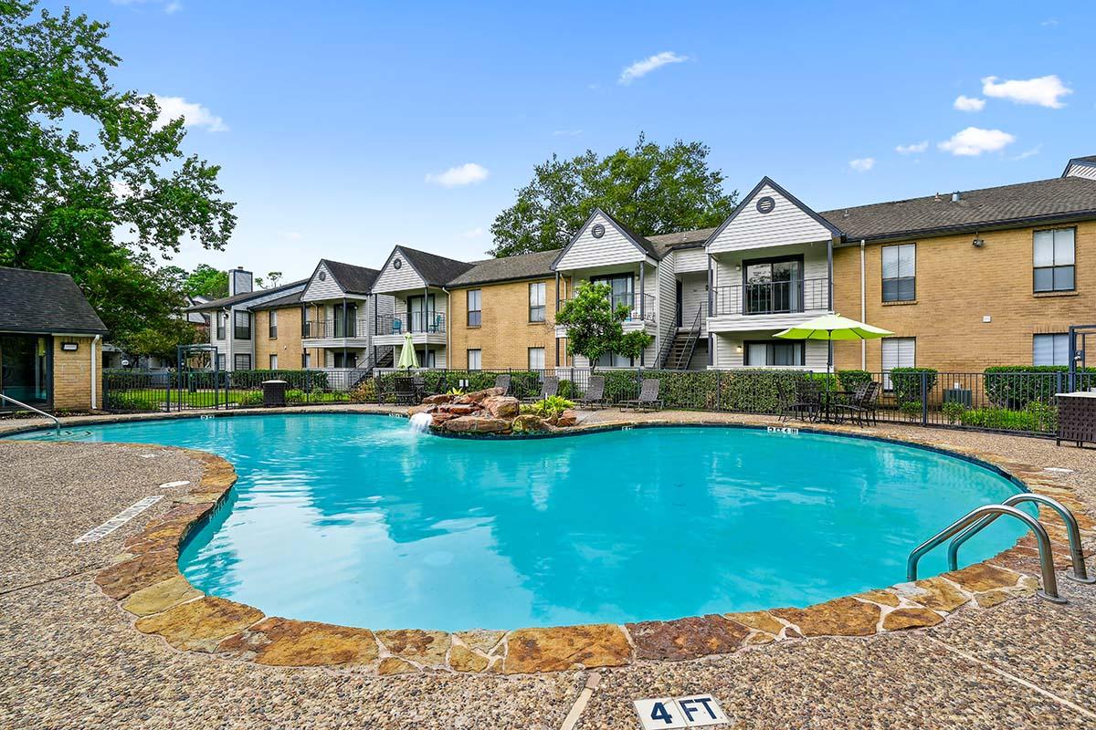 A swimming pool in an apartment complex featuring a rock formation fountain, surrounded by lounge chairs and tables. The backdrop includes two-story buildings with balconies, lush green trees, and a clear blue sky.