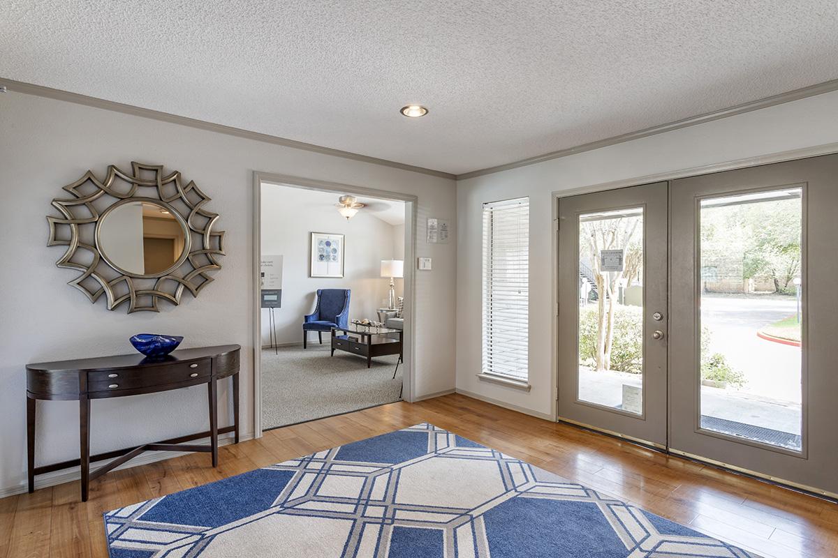 A cozy entrance hall featuring a decorative mirror, a console table with a blue bowl, and a patterned area rug. The space is illuminated by natural light from the large front doors, which open to a view of the outdoors. A living area with a blue armchair is visible through the doorway.