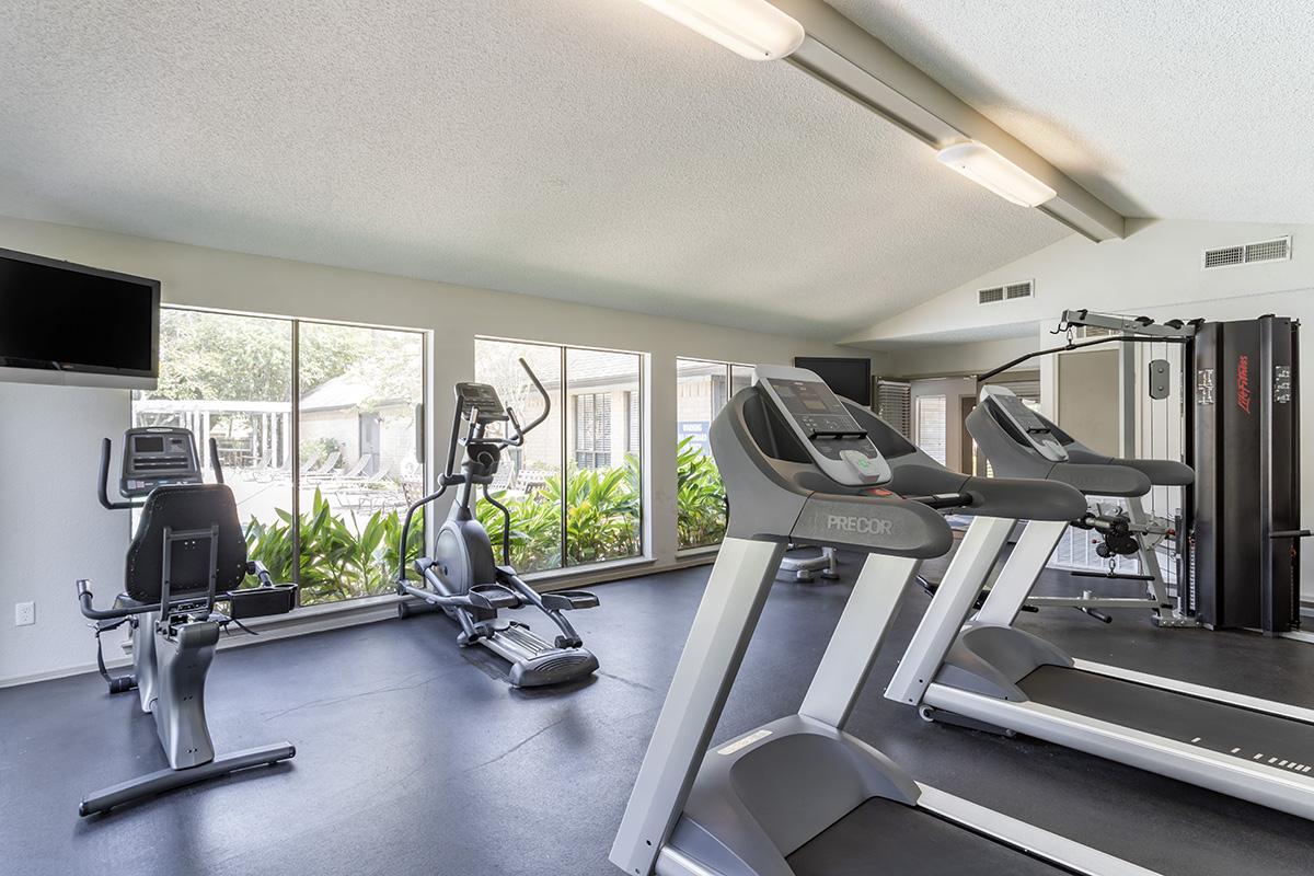 A bright and spacious fitness room featuring several exercise machines, including treadmills, an elliptical, and a multi-gym, all positioned near large windows with views of greenery outside. The room is well-lit, creating an inviting atmosphere for workouts.