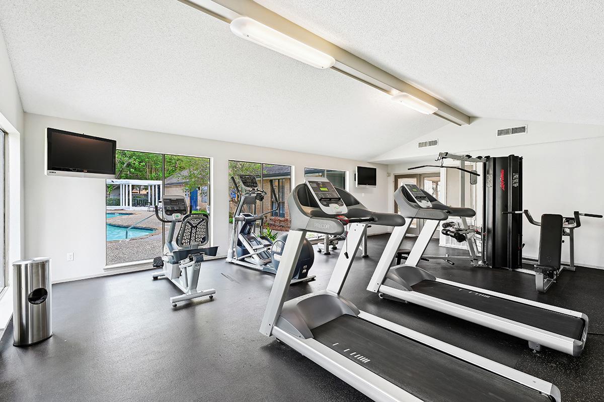 A modern gym with several exercise machines, including treadmills and an elliptical, situated in a bright room with large windows overlooking a swimming pool. There's also a TV mounted on the wall and a weight station, featuring a clean, well-lit space with black flooring.