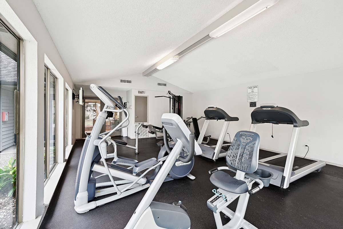 A modern gym interior featuring various exercise equipment, including treadmills, an elliptical machine, and a weight machine. The space is well-lit, with large windows providing natural light and views of the outdoors. The flooring is dark, complementing the sleek design of the equipment.