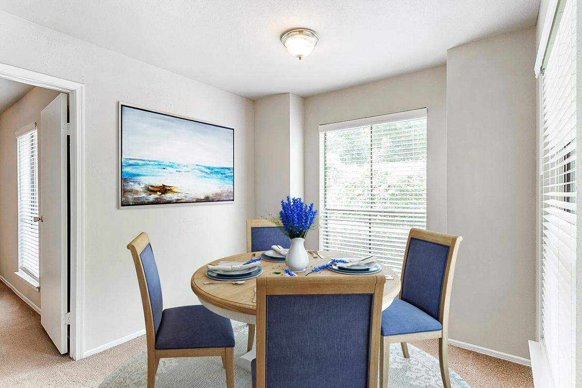 A cozy dining area featuring a round wooden table set with blue plates, a white vase with blue flowers, and four chairs. The walls are light-colored, and a large painting of a seascape hangs on the wall. Natural light streams in through the window, enhancing the inviting atmosphere.