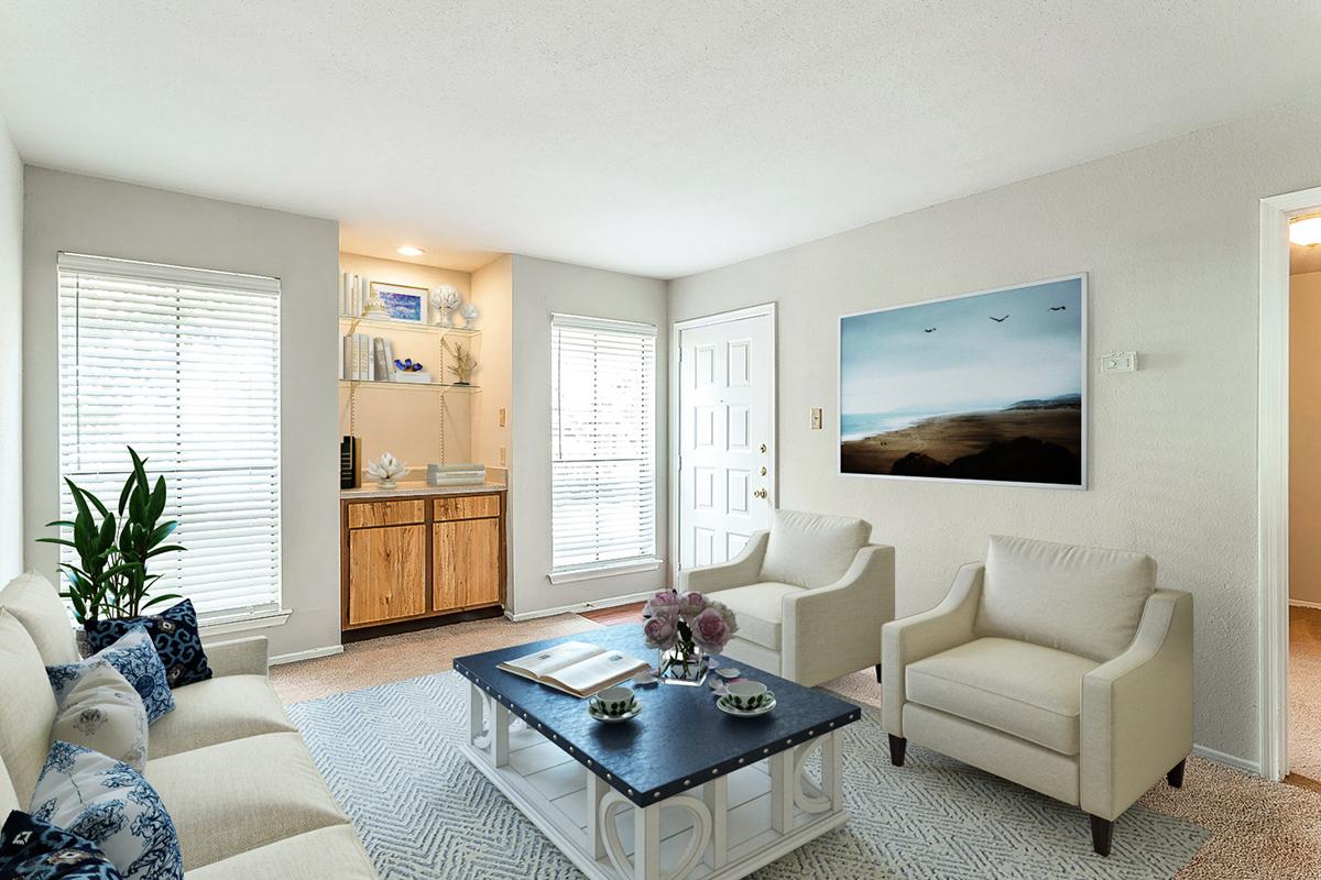 A cozy living room featuring a light-colored sofa with blue patterned cushions, two cream-colored armchairs, and a dark coffee table. There is a wooden cabinet against the wall, a large framed landscape picture, and potted plants. Natural light streams through the windows, creating a warm atmosphere.